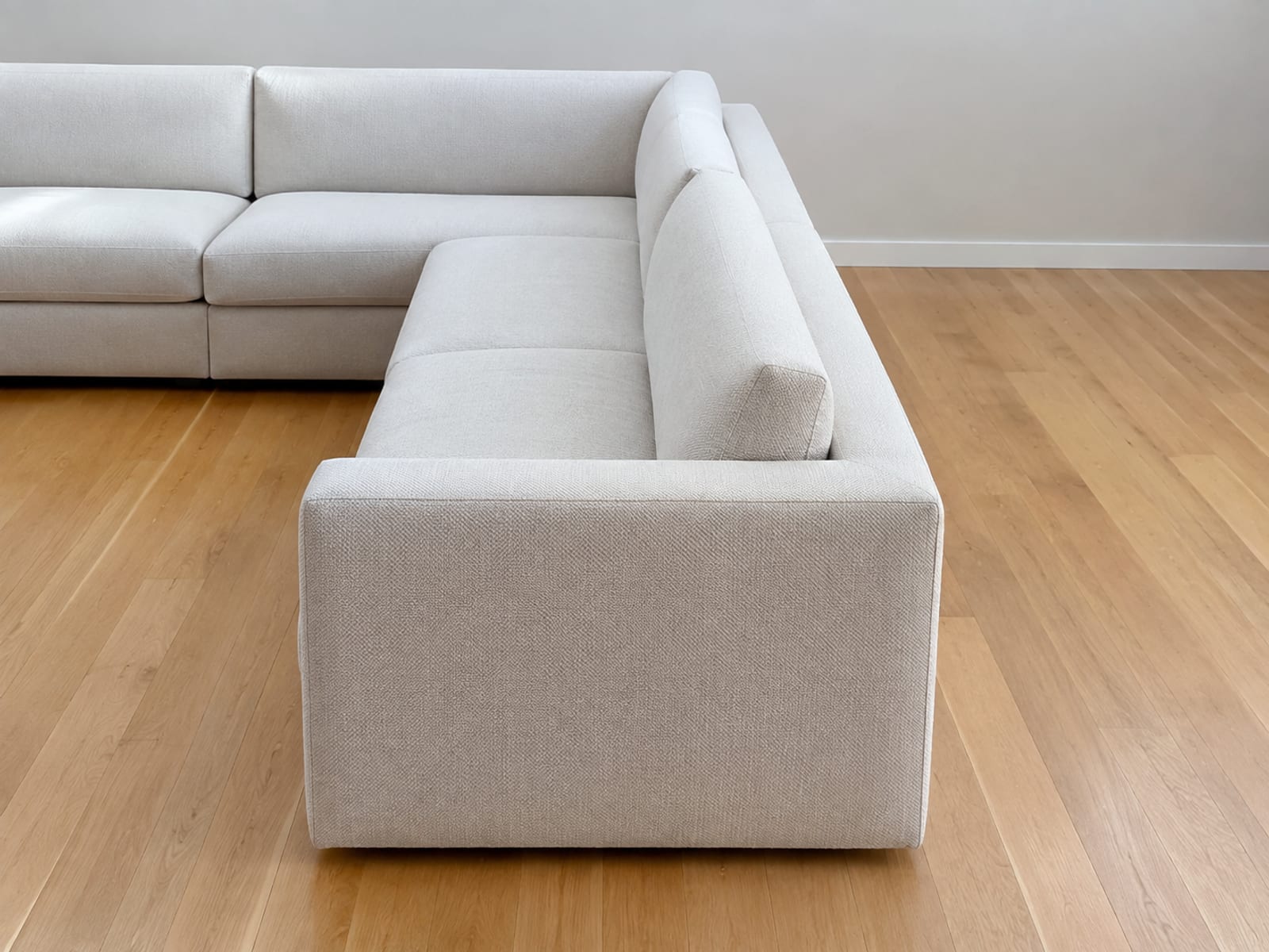 The short return arm of an L-shaped sectional seen along its length from standing height, reaching away from the camera into the room with bare floor on both sides