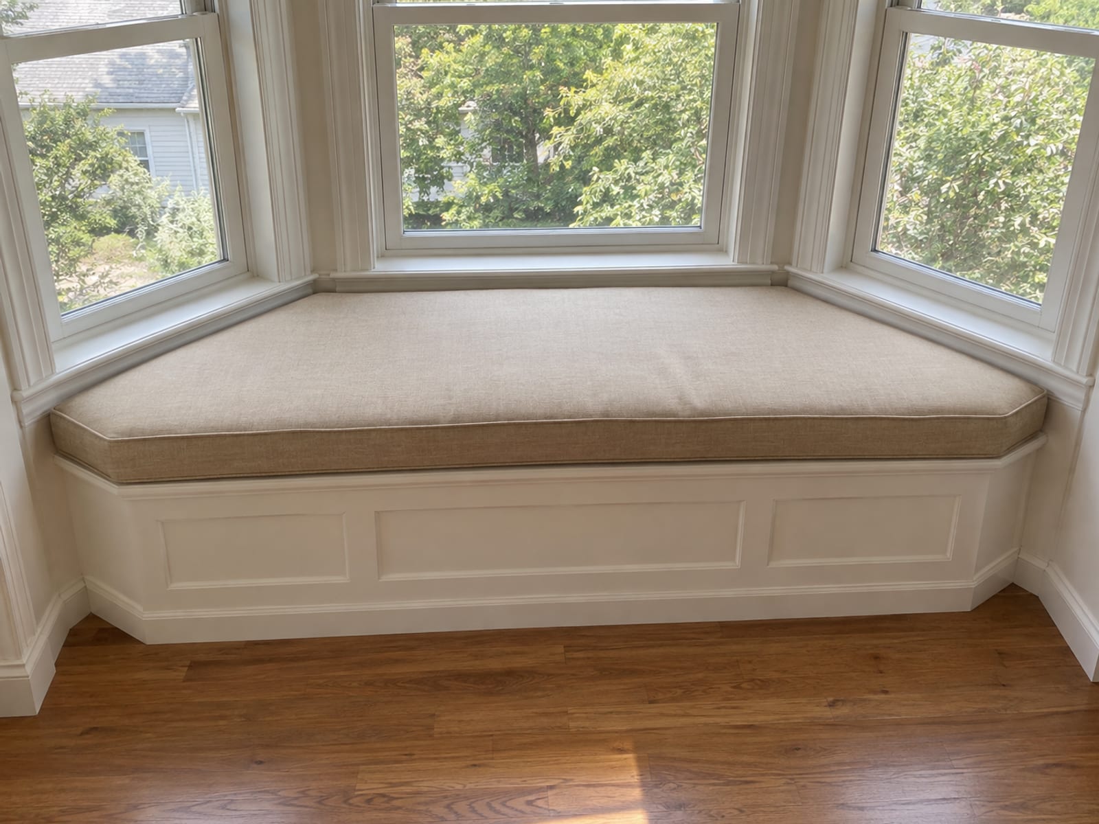 A built-in window seat set into an angled bay window in a daylit room, fitted with one plain upholstered seat cushion cut to follow the bay's angled ends exactly, its sides tapering to meet each angled wall, photographed from standing height slightly above