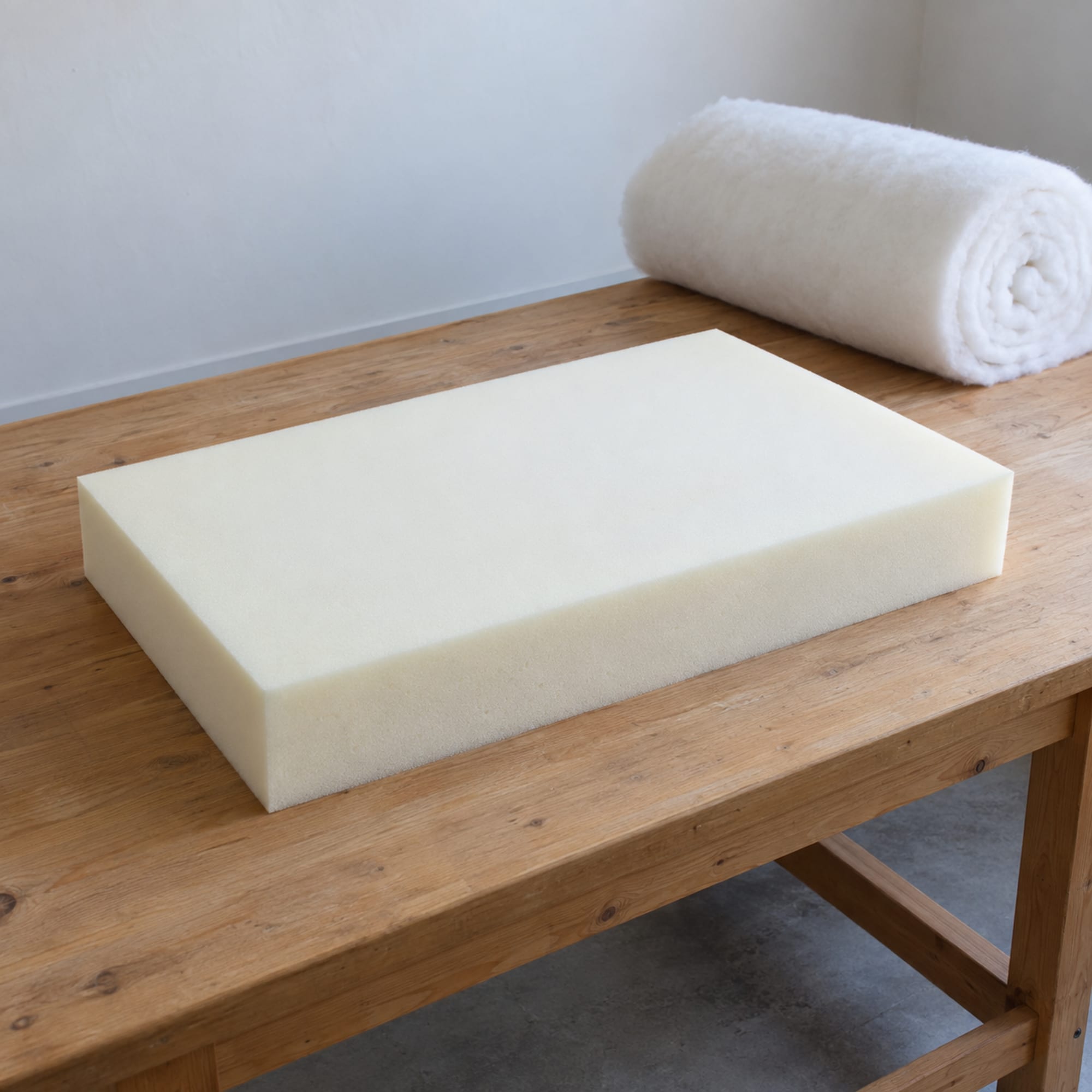 One freshly cut rectangular block of pale cream upholstery foam with crisp square edges lying on a plain wooden workbench in a daylit workroom, with a loose roll of white polyester batting set down at rest at the far end of the bench, photographed from a front three-quarter angle at standing height
