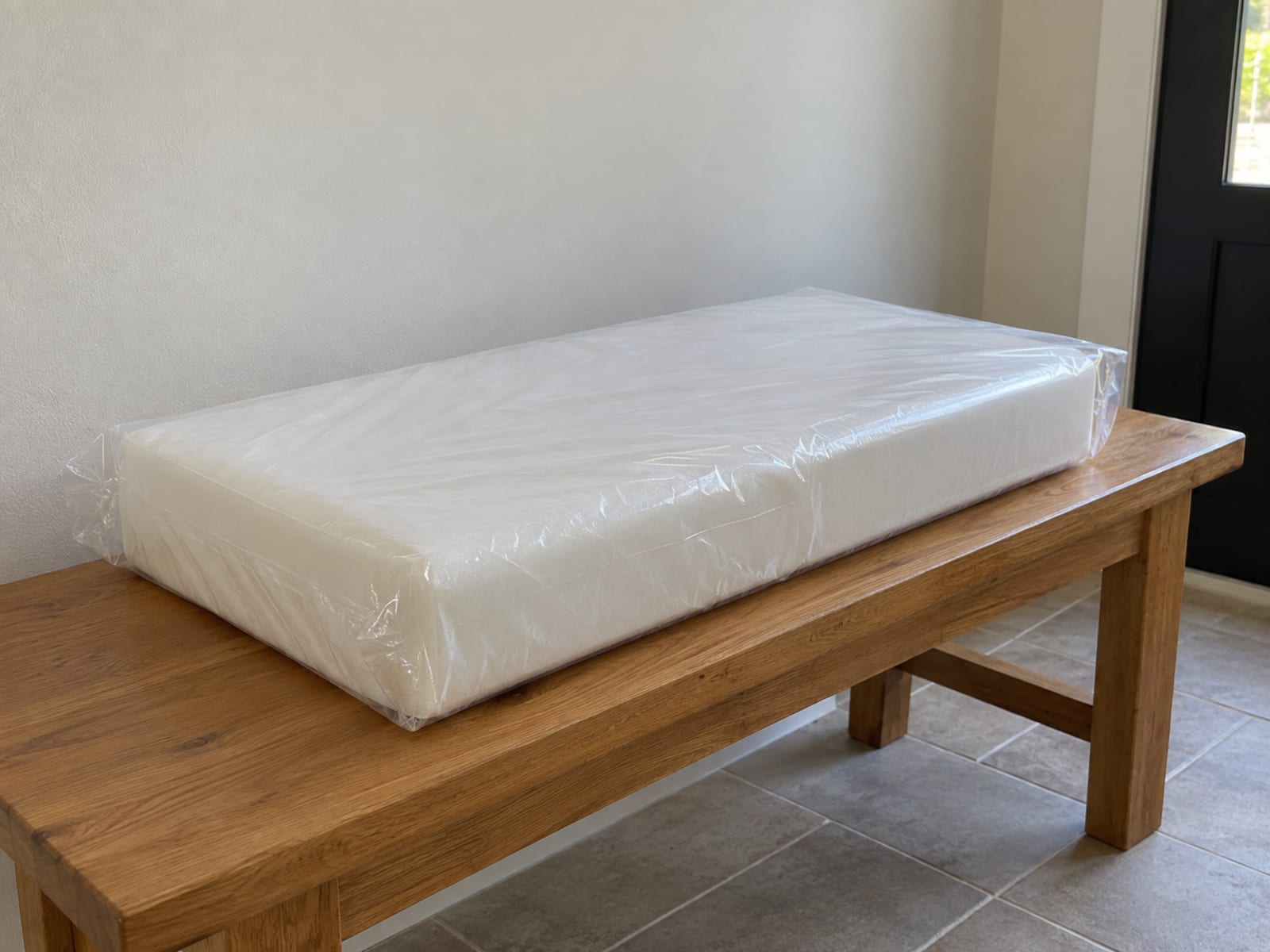 A newly delivered rectangular foam cushion core sealed in clear, completely unprinted plastic film lying flat on a plain wooden bench in a daylit entry hall, photographed from standing height at a front three-quarter angle