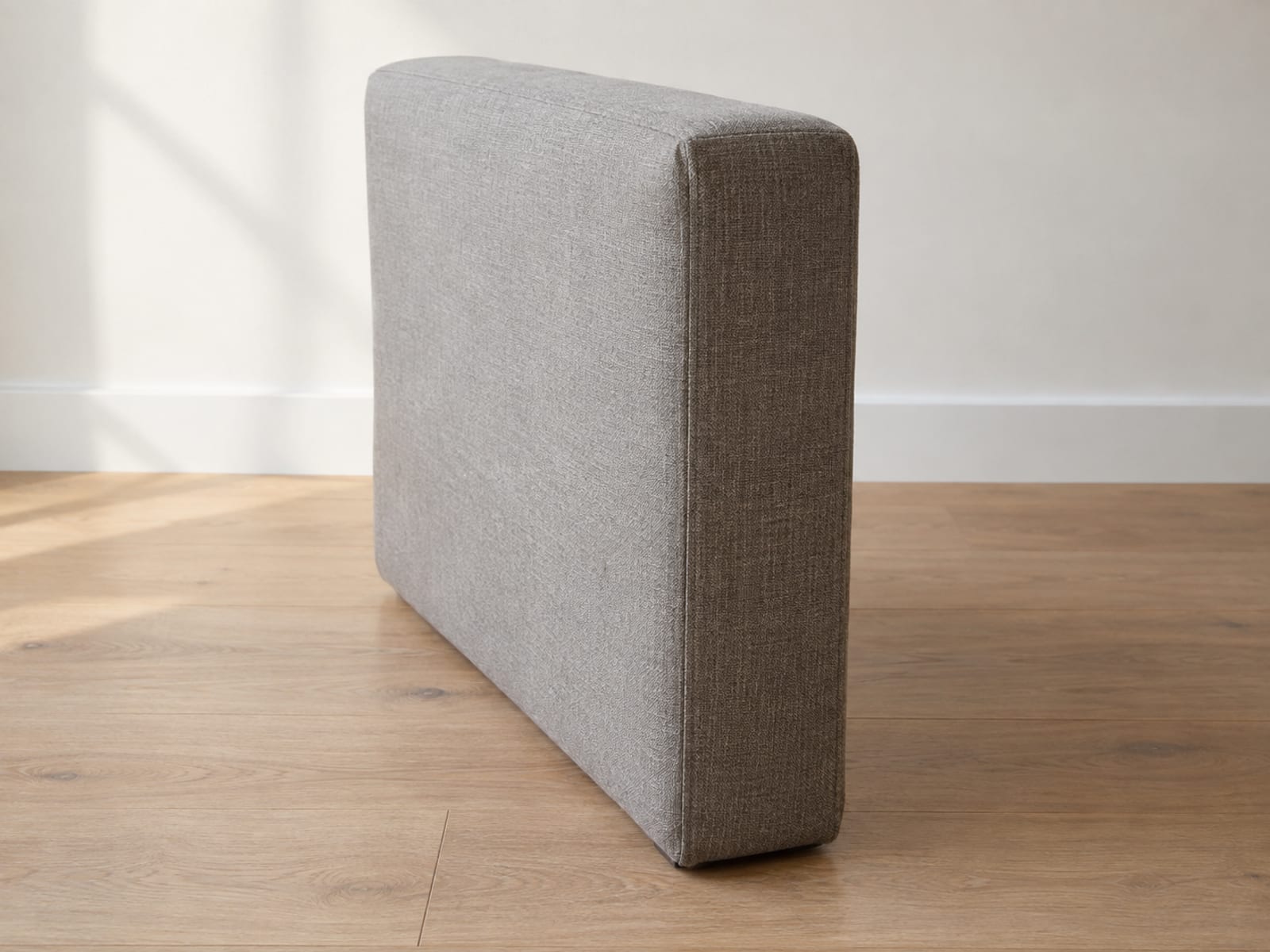 One separate upholstered sofa arm section, a boxy panel in a warm gray woven cover, standing upright on its own on bare oak floorboards in front of a plain wall in an empty daylit room, photographed square on from the side of the arm at standing height so it reads as a spare part made to fit a sofa rather than a piece of furniture
