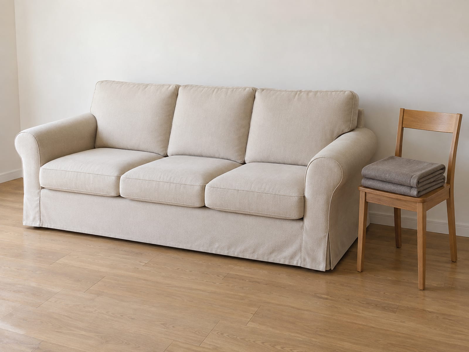 A three-seat sofa wearing a pale beige cover in a daylit living room, and beside its arm a plain wooden dining chair with a second, complete spare sofa cover in a darker warm gray folded into one neat squared bundle on its seat, photographed from standing height at a three-quarter angle so the sofa and the folded spare beside it read together
