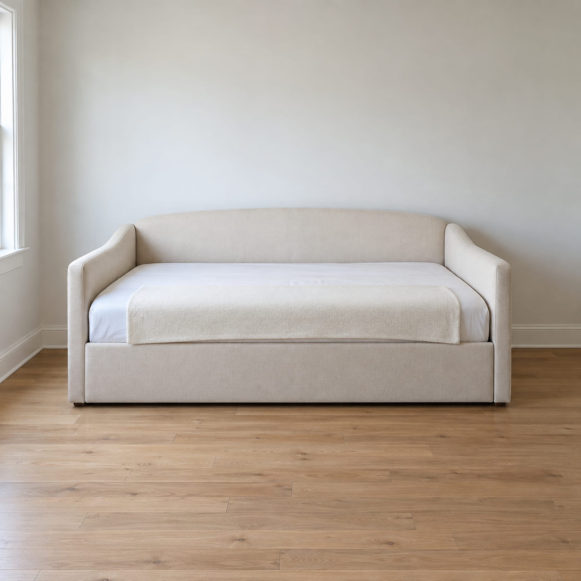 An upholstered twin daybed with a plain padded back and two padded end arms, made up plainly with a single pale fitted sheet and one folded pale blanket laid across its foot, set against the wall of a small daylit spare room, photographed square on from the front at standing height