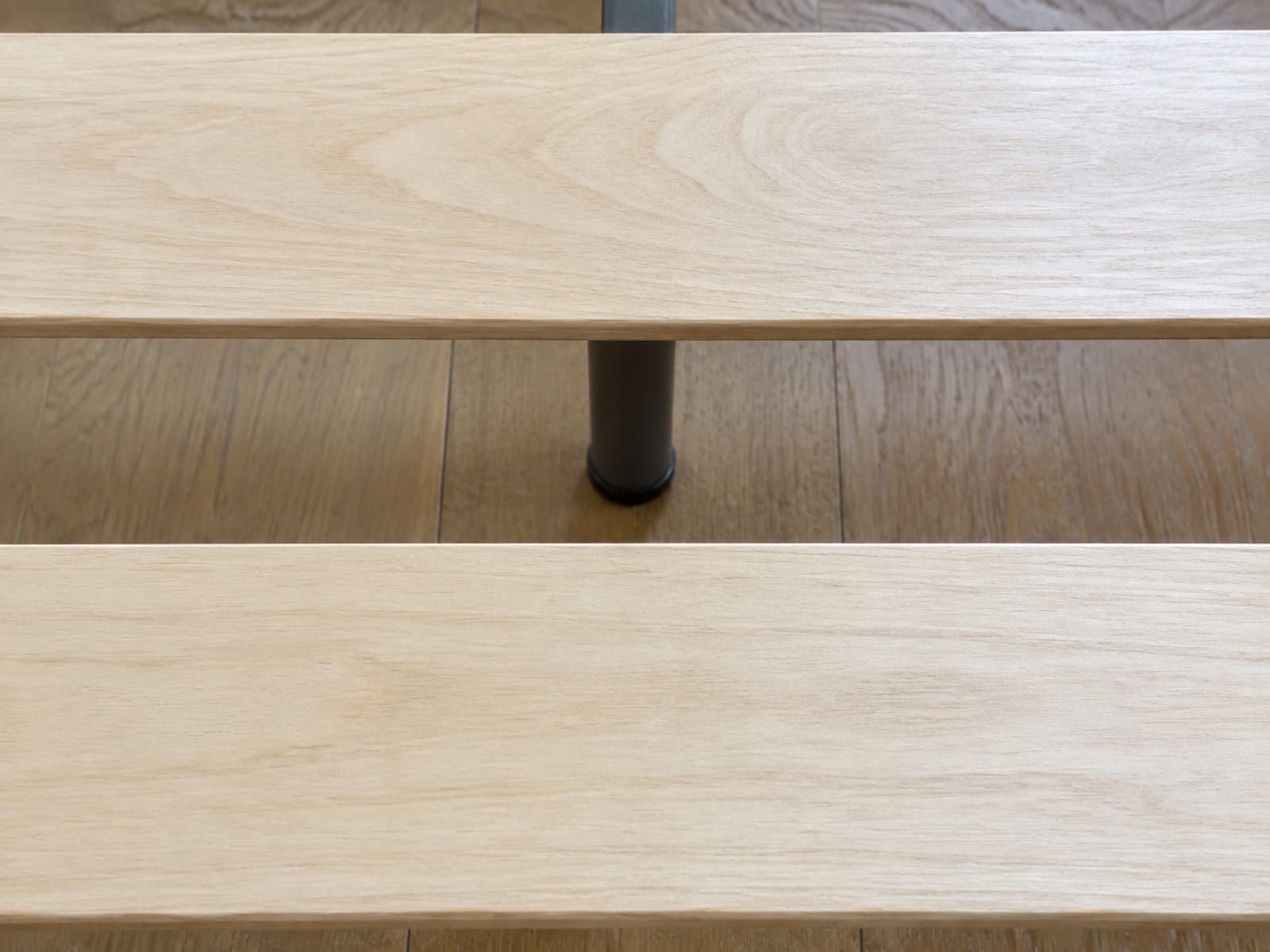 An extremely close view from above of two flat wooden daybed slats running across the frame with the even gap between them in the middle of the picture, and through that gap the plain metal center support rail and one of its short feet visible below, standing on bare floorboards