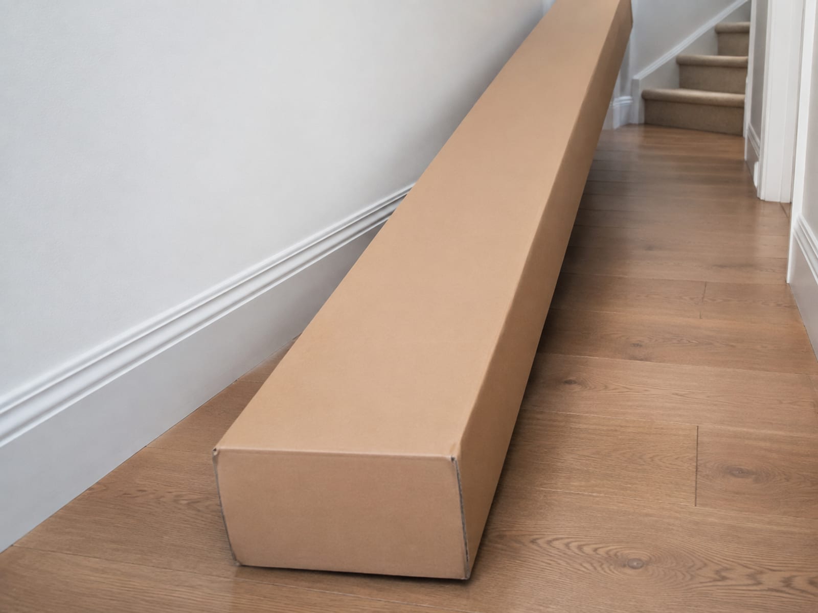One very long plain unmarked carton lying flat on the bare floorboards of a narrow hallway, running out of frame, with the bottom of a staircase and its turn visible beyond