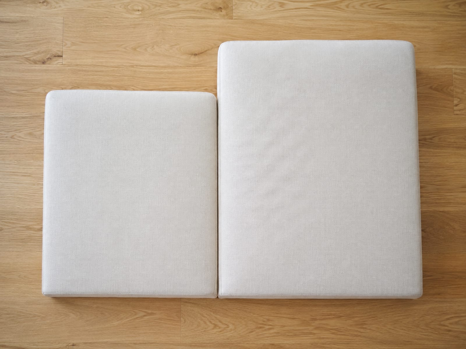 Two plain pale seat cushions of clearly different front-to-back depth lying flat side by side on bare floorboards, their front edges aligned so the difference shows at the back
