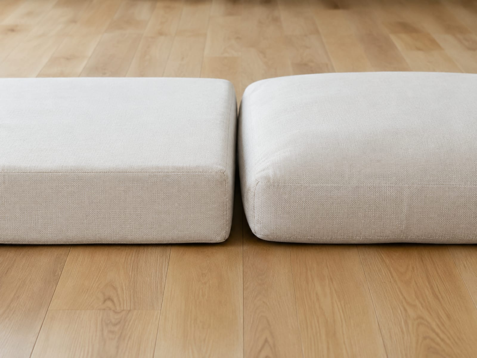 Two seat cushions of the same footprint side by side on floorboards seen from floor level, the left one square-edged and flat-topped, the right one softer and rounded where a fiber wrap fills the cover