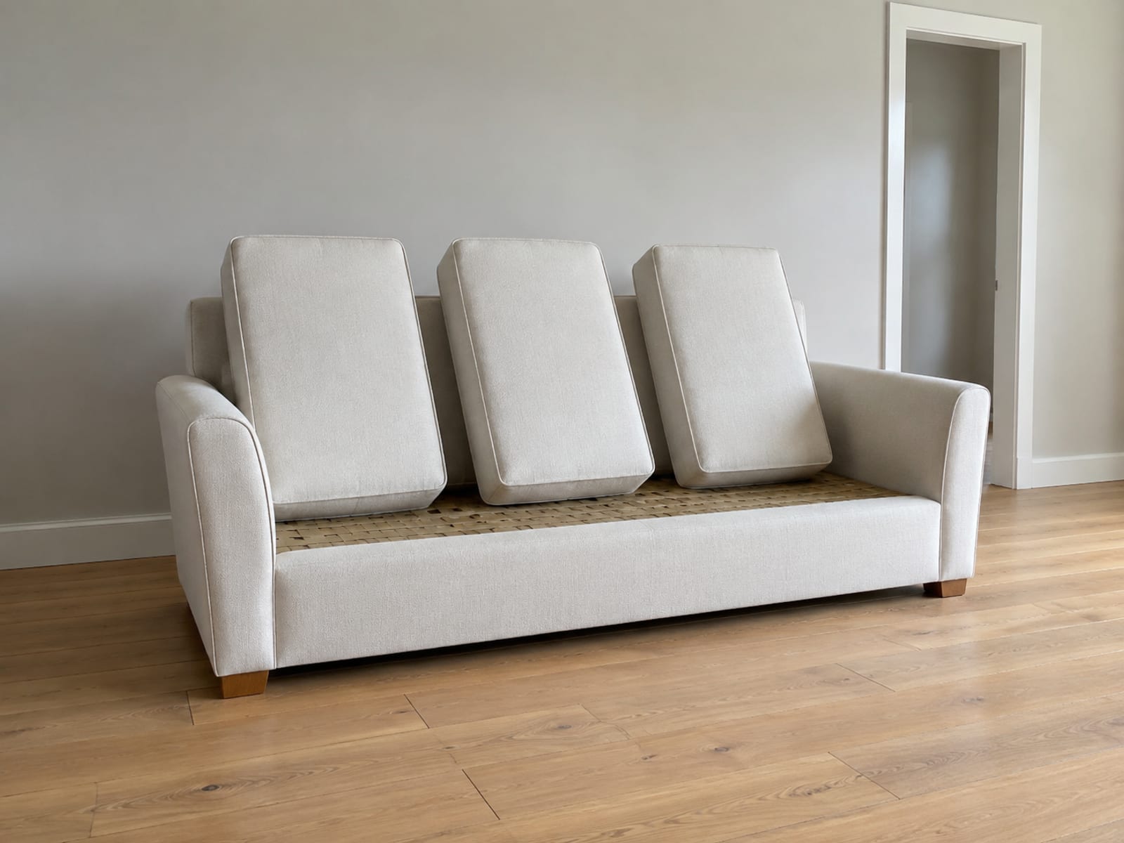 A pale three-seat sofa alone in an empty daylit room with its seat cushions stood on their edges along the seat