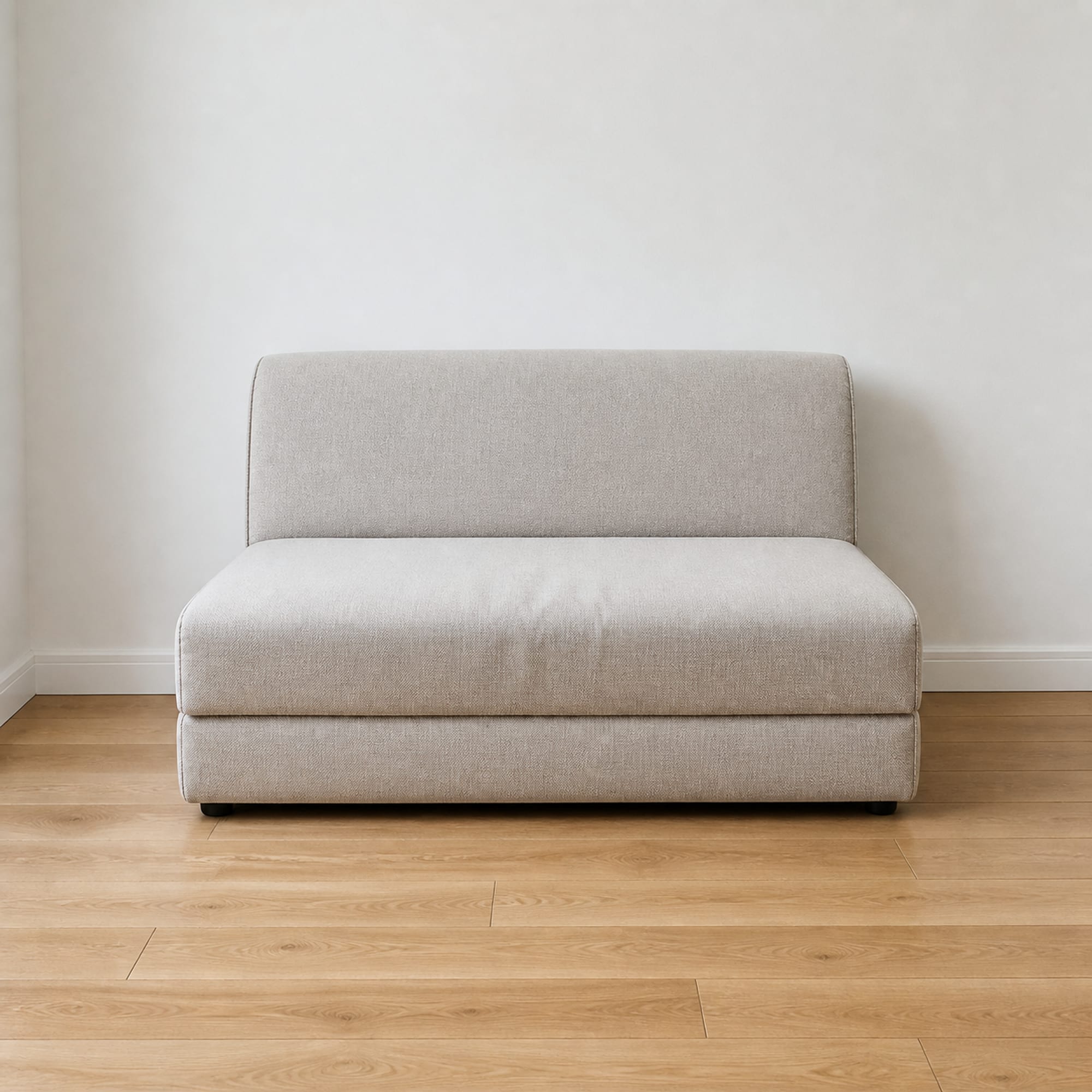 A compact convertible sofa in its seating position in a small bare room, photographed at seat height so its low seat and short back read the way they would to somebody about to sit down
