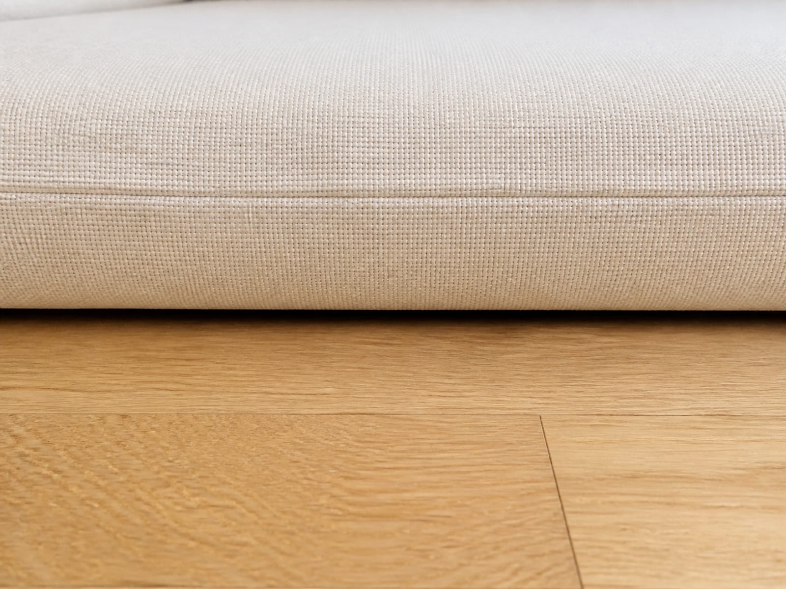 An extremely close view from floor level of the front edge of a convertible sofa seat cushion and the bare oak floorboards immediately below it