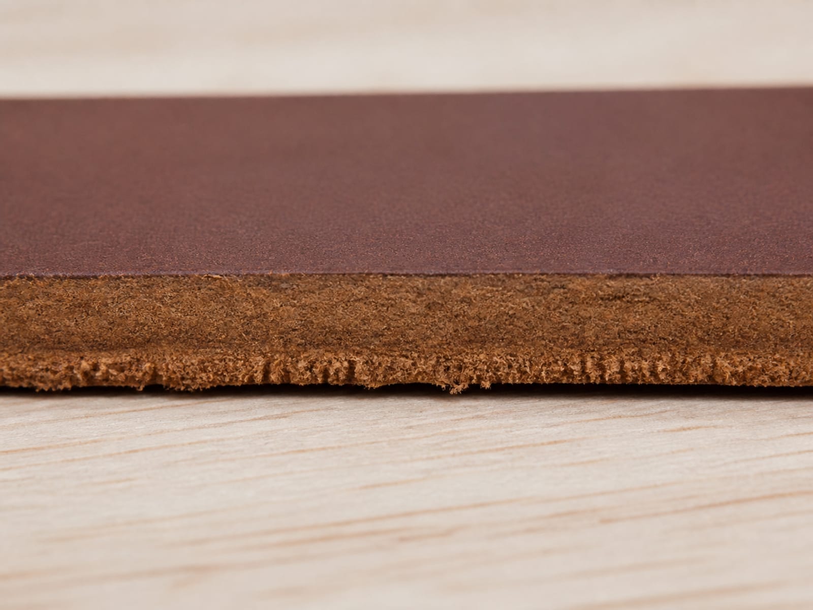 An extremely close end-on view of the raw cut edge of a thick piece of mid-brown leather, the finished face above and the softer fibrous underside beneath