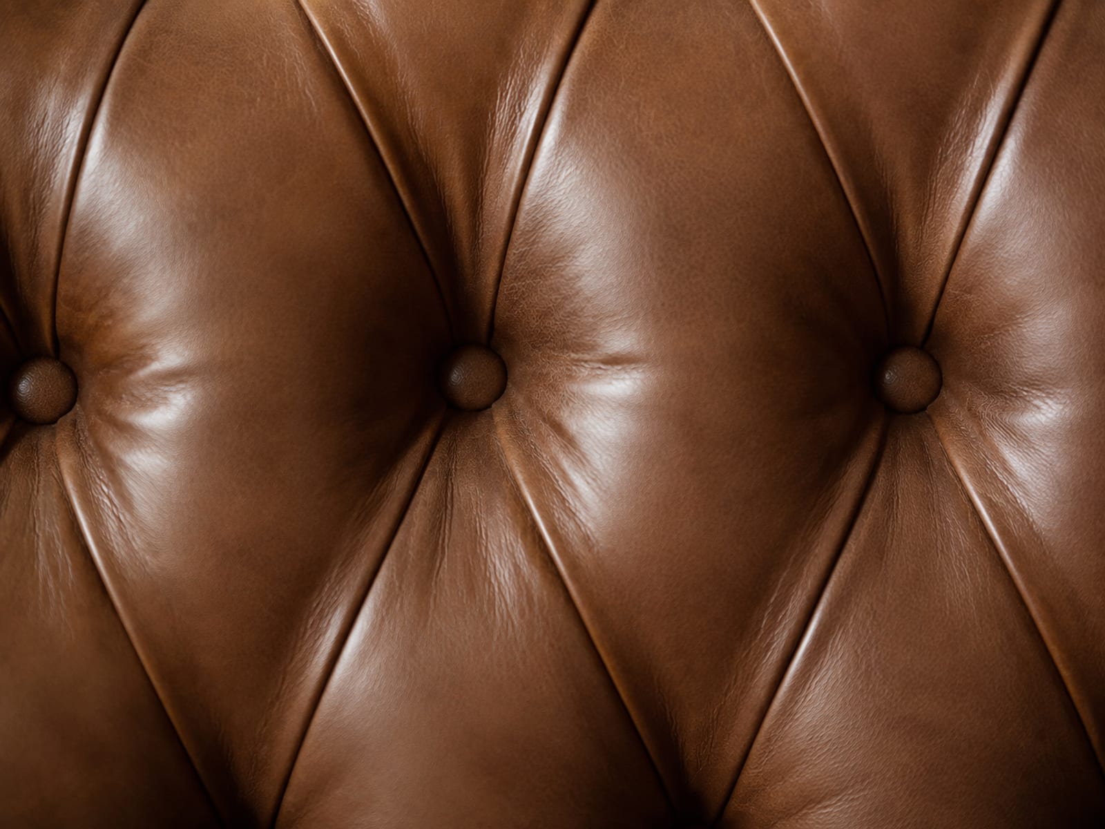 A close view onto the tufted back of a mid-brown leather sofa, the hide drawn into deep faceted folds radiating from each covered button