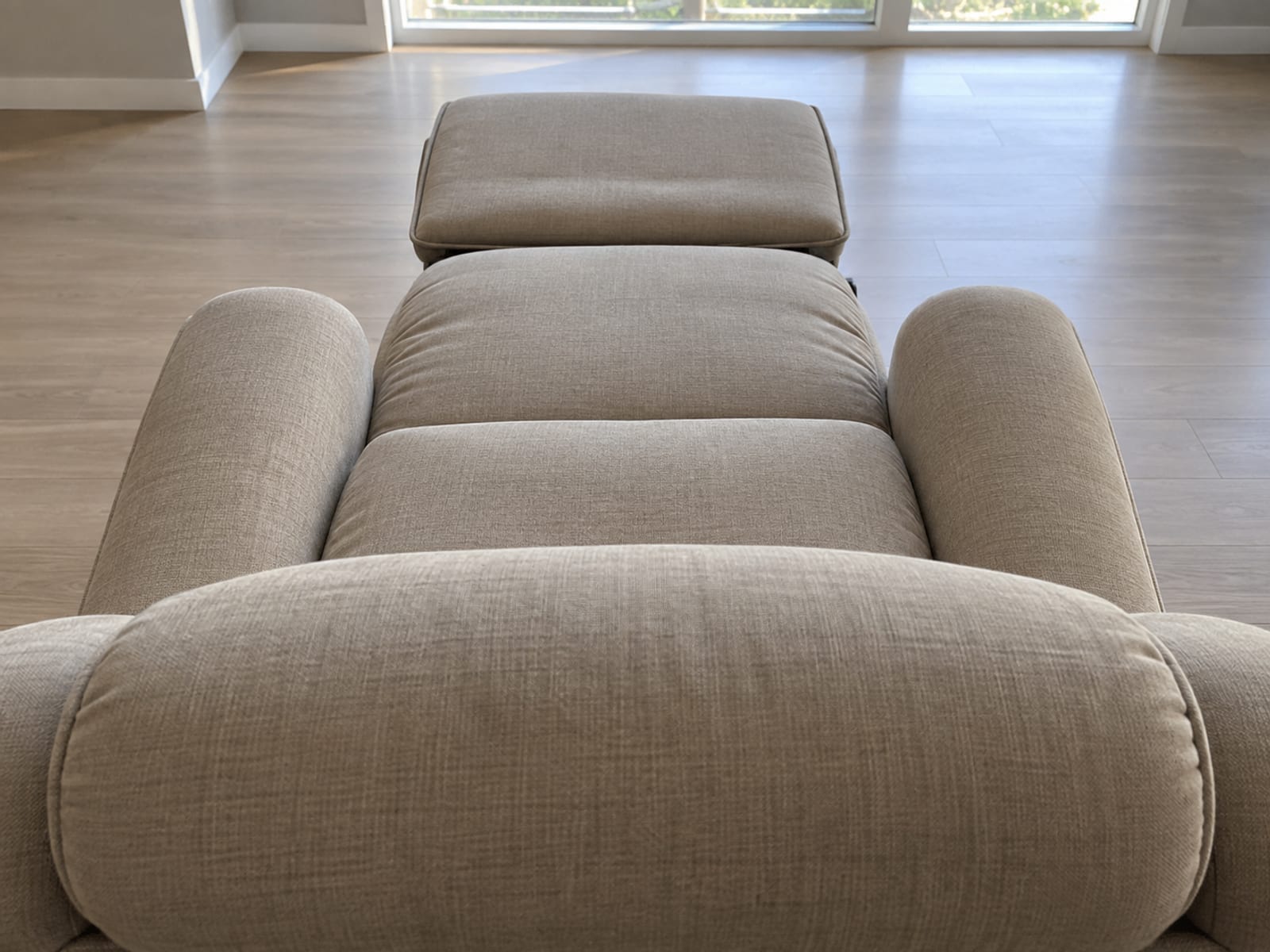 An upholstered lift chair reclined almost fully flat in an empty daylit room, photographed from a raised position just behind and above its headrest, looking down along the whole length of the reclined chair toward the raised footrest at the far end