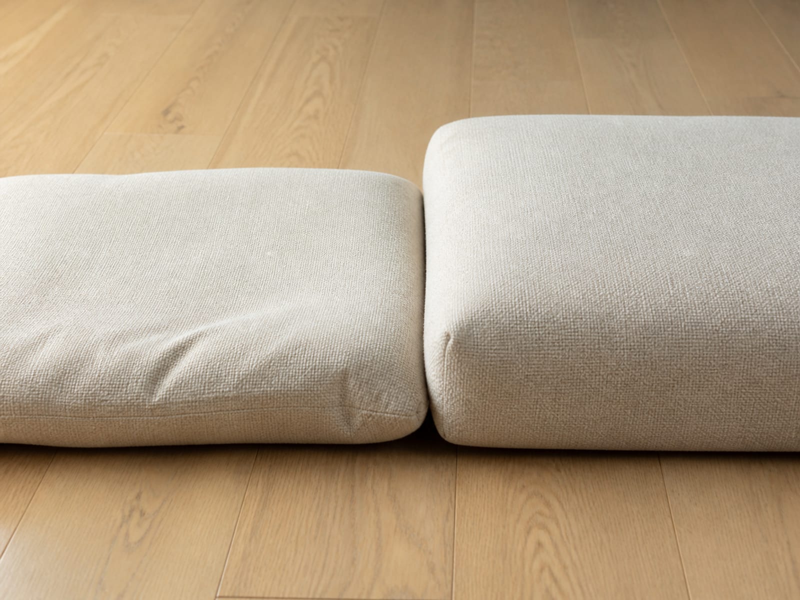 Two seat cushions of identical size in the same pale cover lying side by side on bare floorboards under one raking light, the left settling lower and rounder than the right