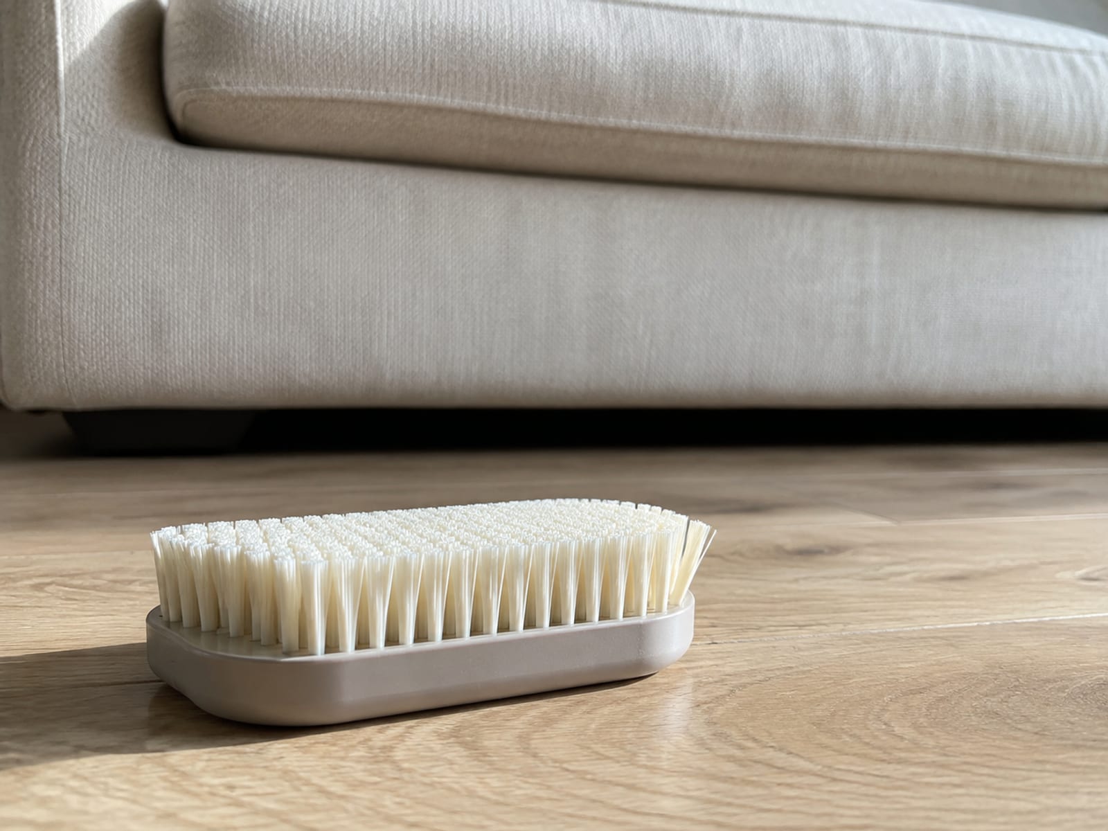 A plain brush with dense pale bristles lying bristles-up on floorboards beside a sofa, its bristles empty and the seat cushion above swept clear