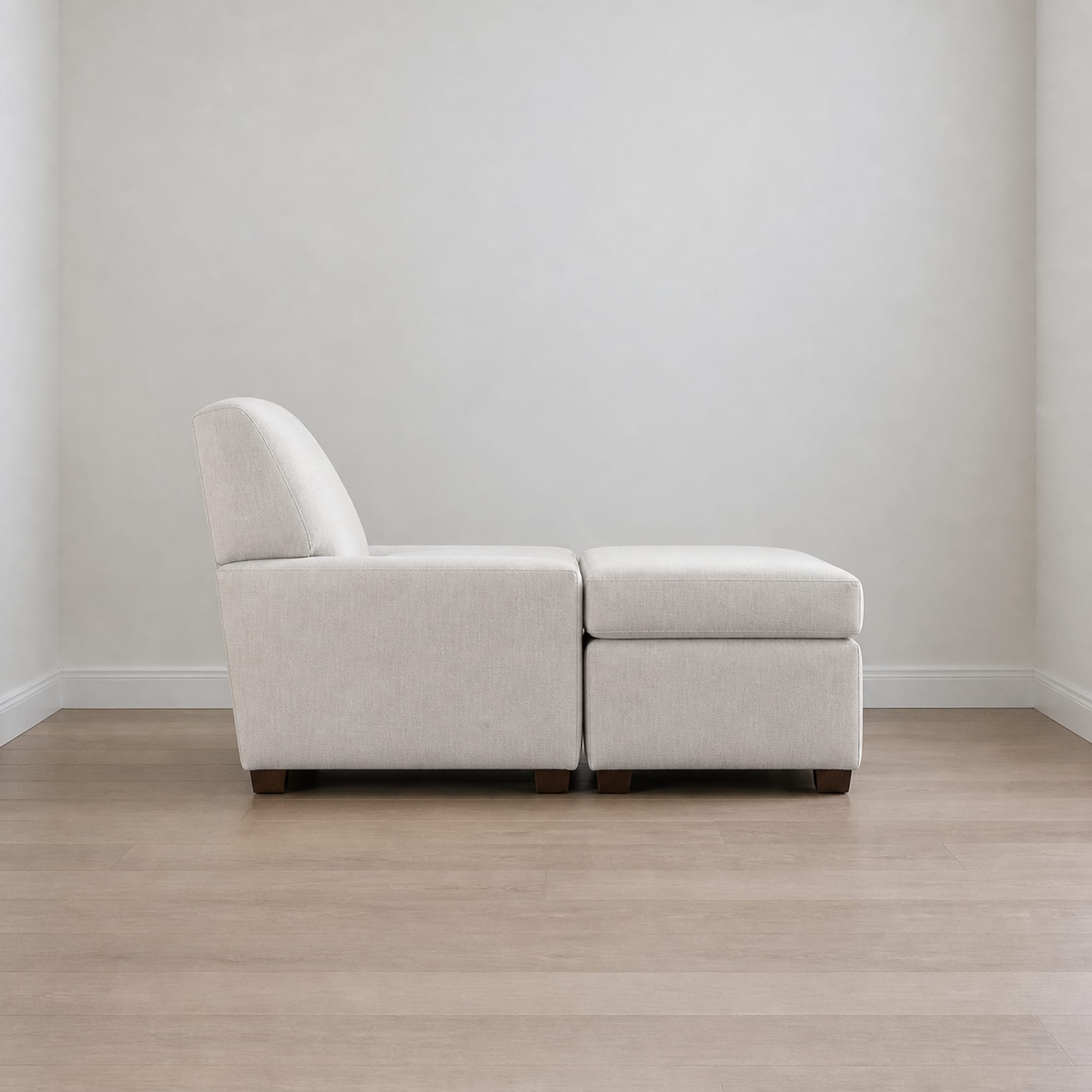 An upholstered armchair with a matching footstool directly in front of it in a bare corner, seen square from the side so the seat and the stool read as one continuous surface
