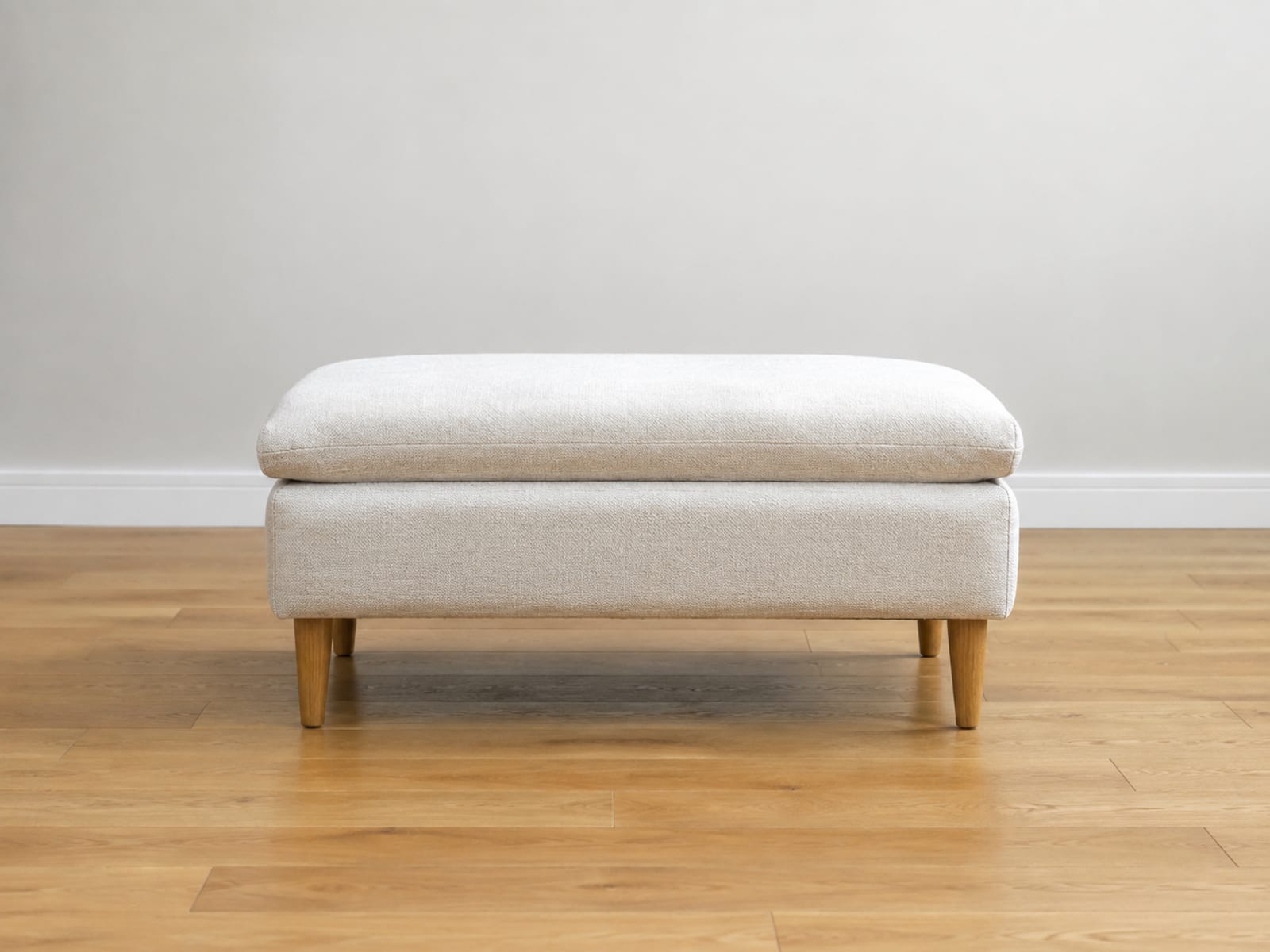 One upholstered footstool standing by itself on bare oak floorboards in an empty room, seen square from the side at its own height