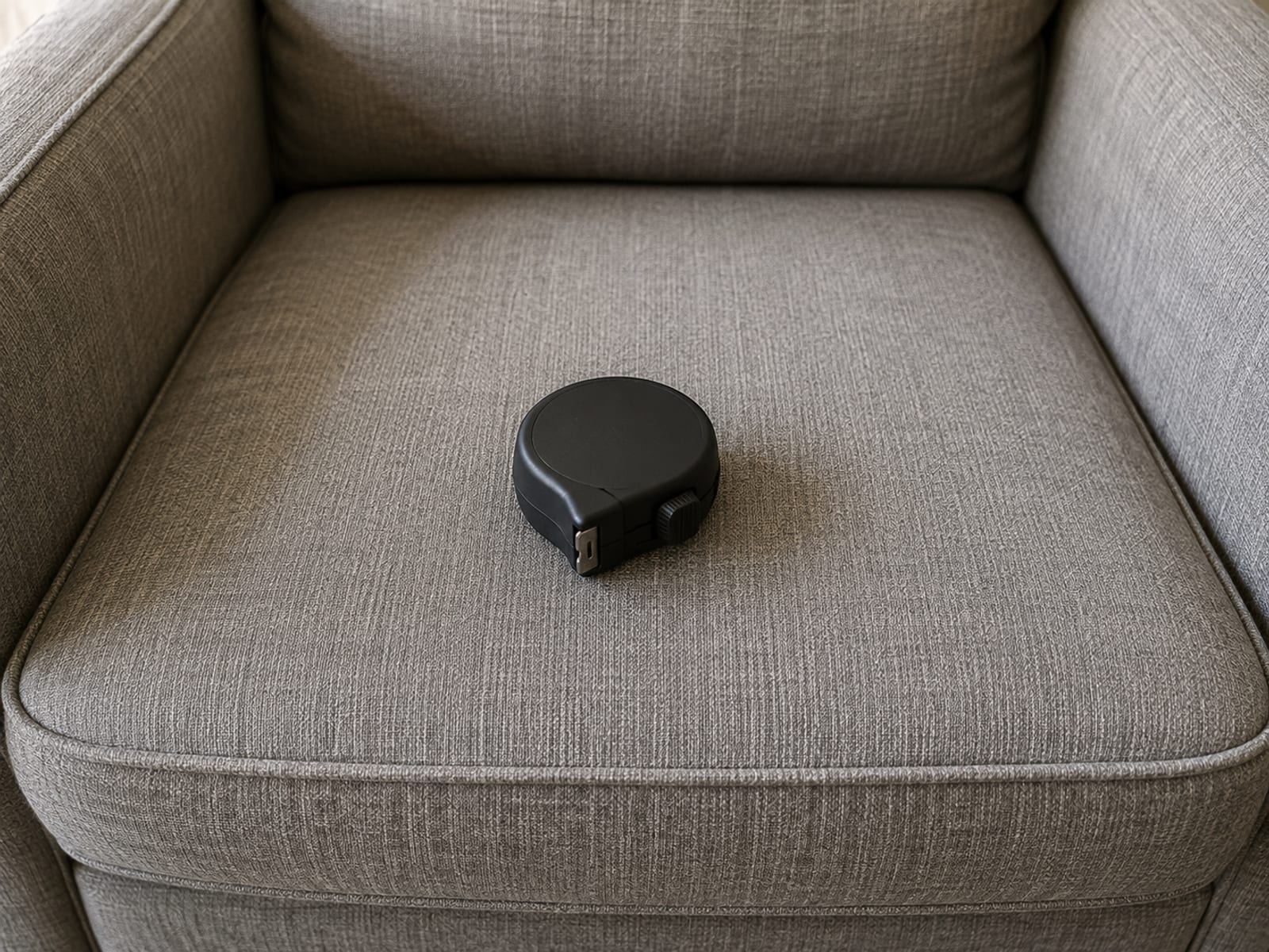 A small retractable tape measure, closed and coiled in its case, resting on the bare seat of an uncovered recliner