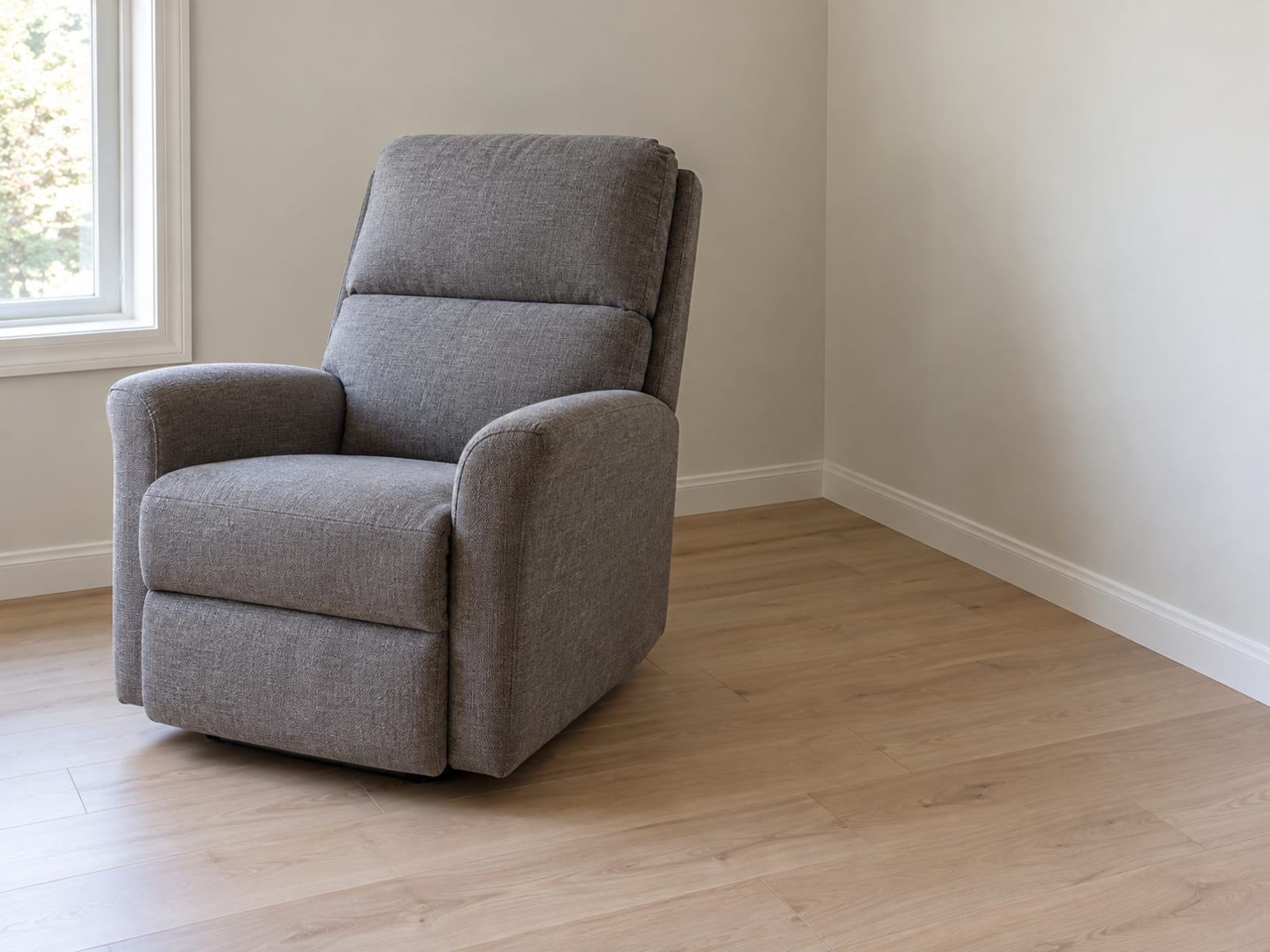 An upholstered wall-hugger recliner in a mid-gray woven fabric, closed and upright, set diagonally across the corner of a plain daylit living room so an open triangle of bare floorboards lies between the back of the chair and the two walls meeting behind it, photographed from across the room at standing height at a gentle downward angle
