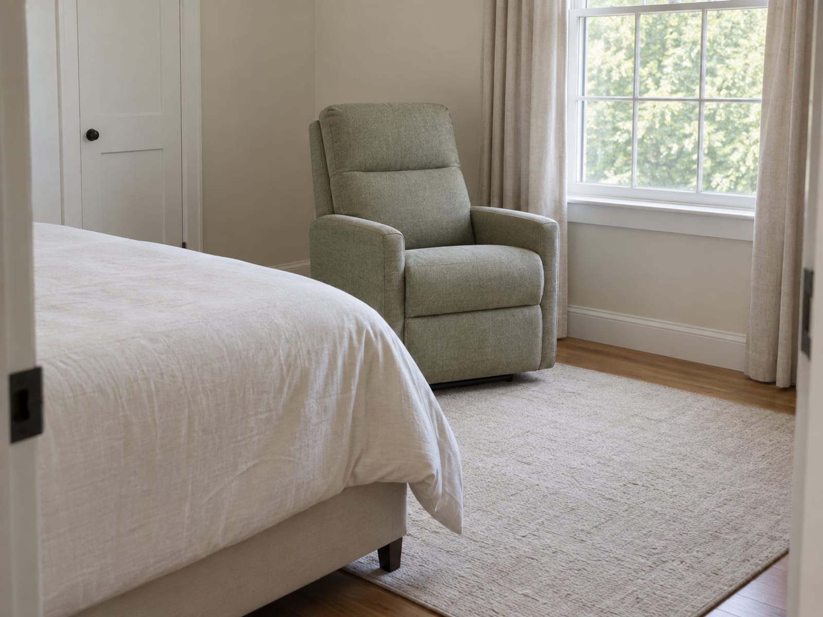 A compact upholstered recliner with slim track arms, closed and upright, standing in the space between the foot of a plainly made bed and a window in a small daylit bedroom, photographed from the bedroom doorway at standing height so the corner of the bed in the near foreground and the chair beyond it read as one small room