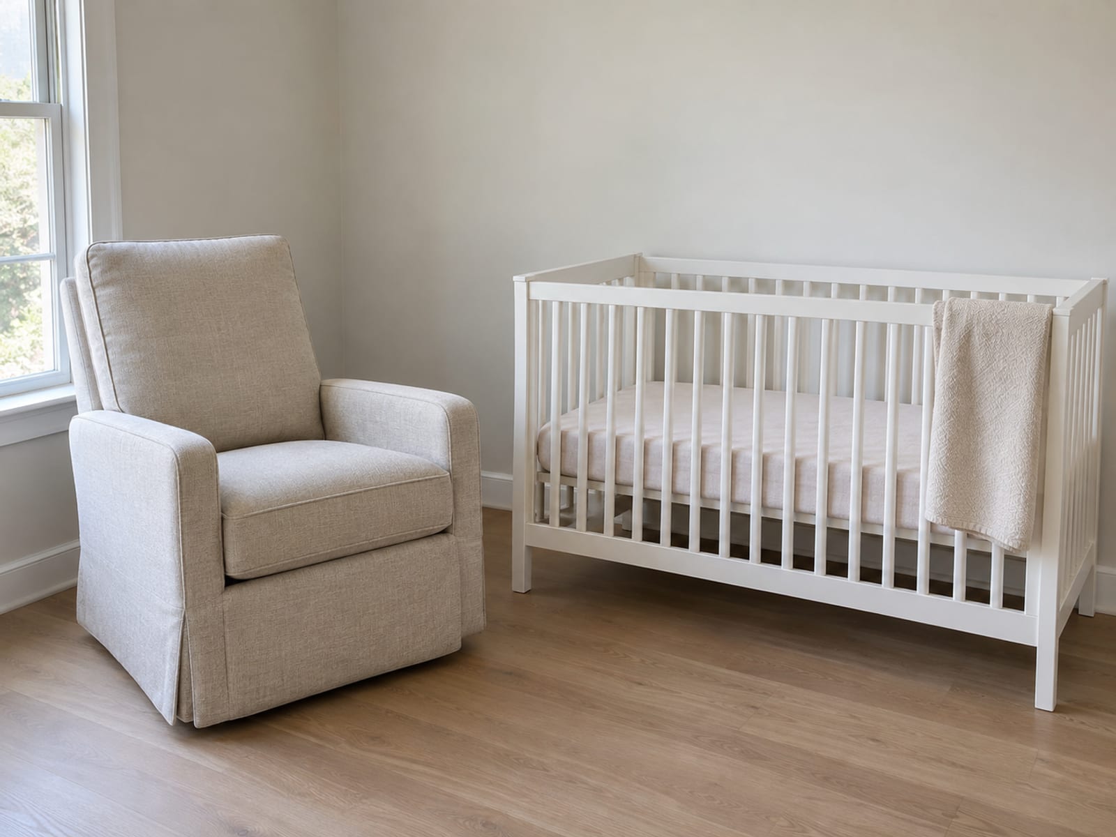 An upholstered swivel glider recliner in a pale oatmeal fabric, closed and upright and angled toward the room, standing in the corner of a small daylit nursery beside a plain white-painted wooden crib against the adjacent wall, with a soft plain folded blanket hung over the crib rail, photographed from standing height at a front three-quarter angle