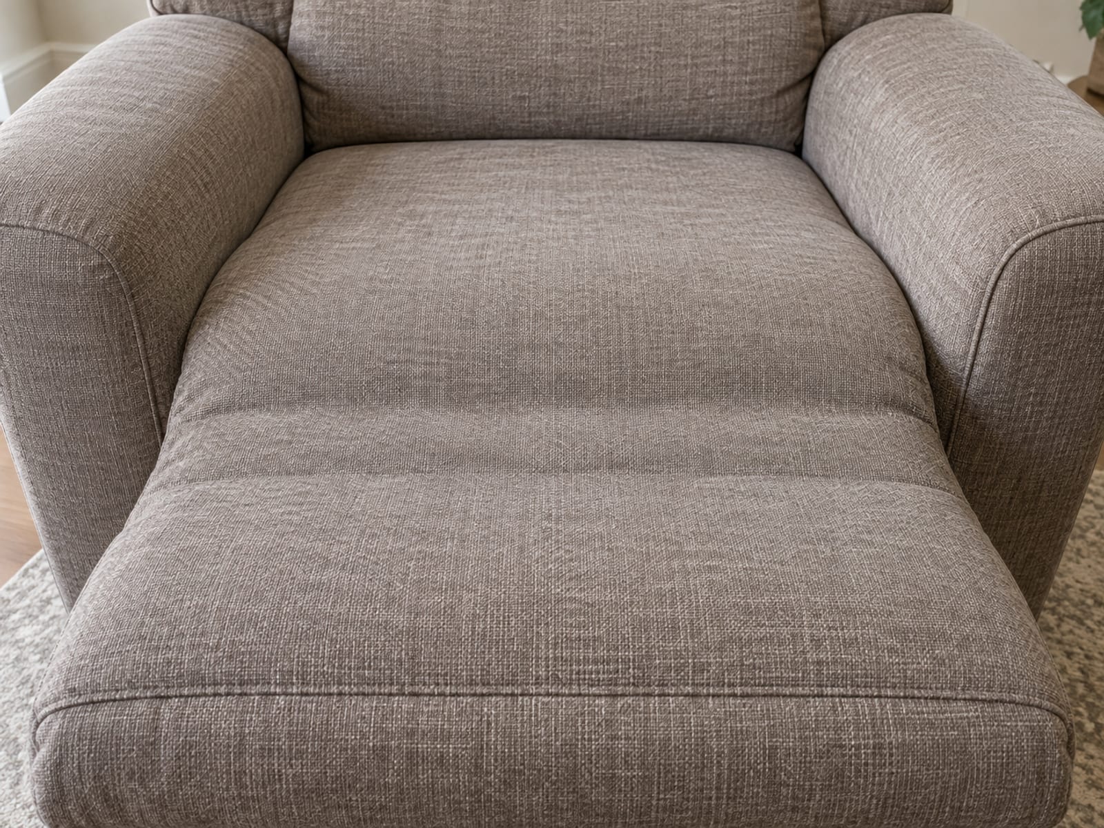 A close view from standing height looking down at a slight angle onto the middle of a reclined upholstered recliner, where a continuous padded chaise panel runs from the front of the seat cushion out over the join and onto the raised footrest as one unbroken upholstered surface, framed so the seat, the padded bridge and the footrest fill the picture from side to side