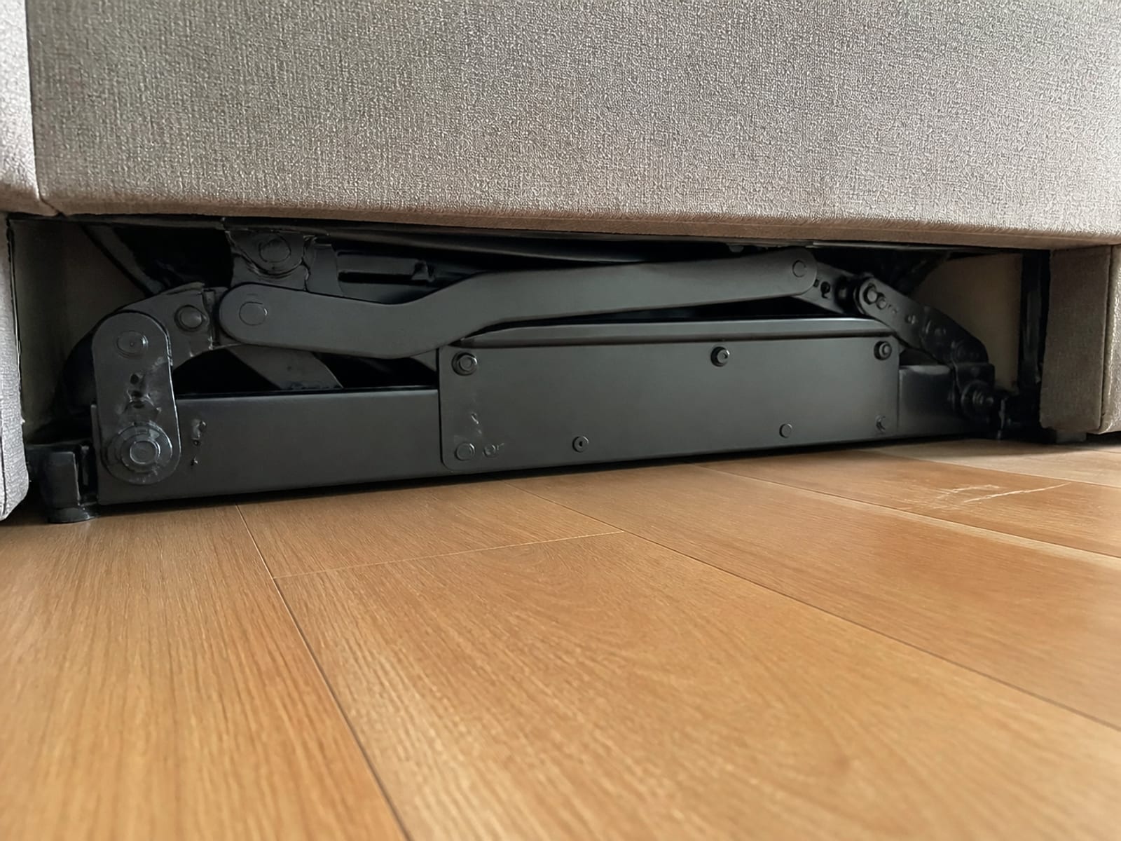 A low view from floor level in under the front edge of a reclining sofa, the folded steel mechanism housing visible as a boxed structure of plates and pivots