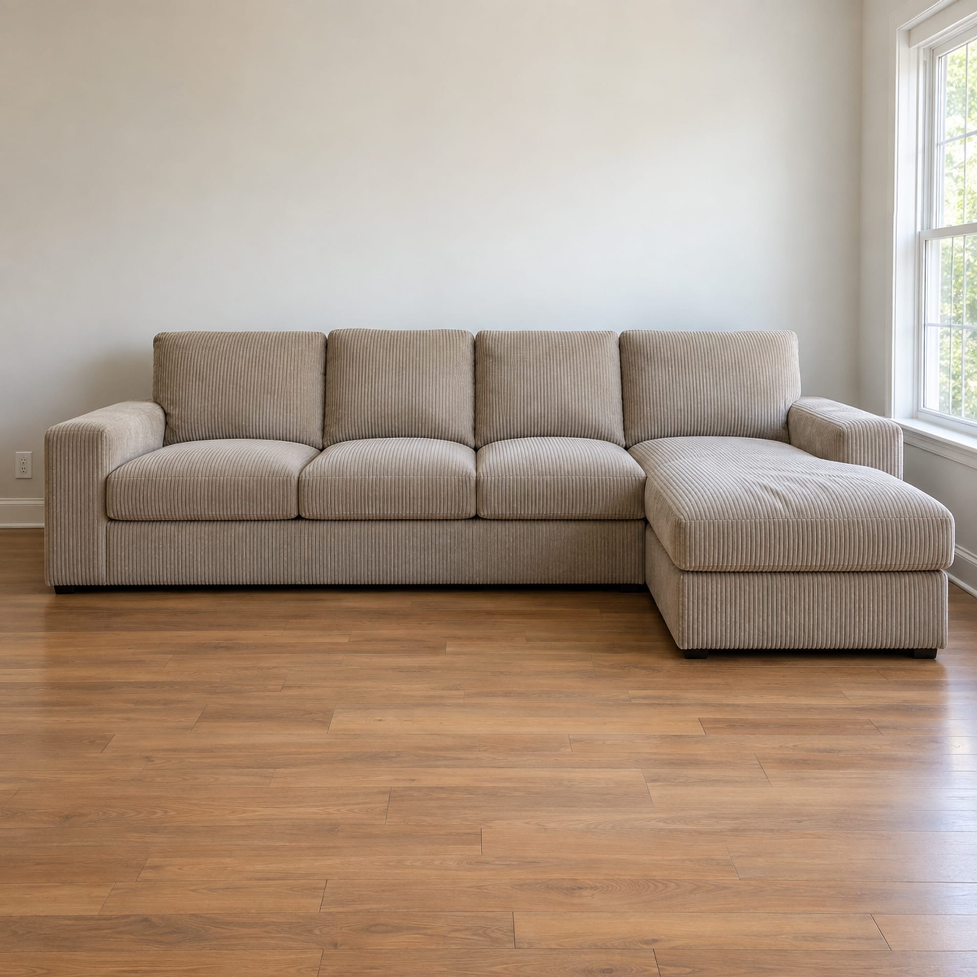 A modest four-seat L-shaped sectional in a warm putty corduroy standing against the long wall of a small first-apartment living room, photographed square on from the front at standing height, the chaise reaching toward the camera on the right and the straight run flat against the wall behind