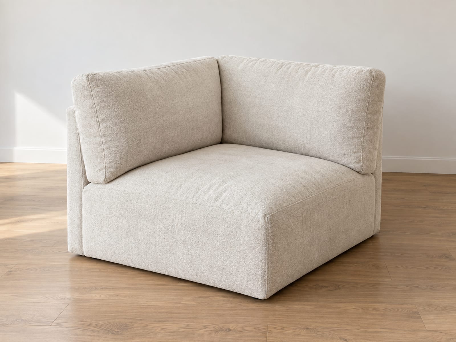 The square corner unit of a modular sectional standing entirely alone on bare oak floorboards in an empty daylit room, photographed from a front three-quarter angle at standing height, its two upholstered back pieces meeting at a right angle