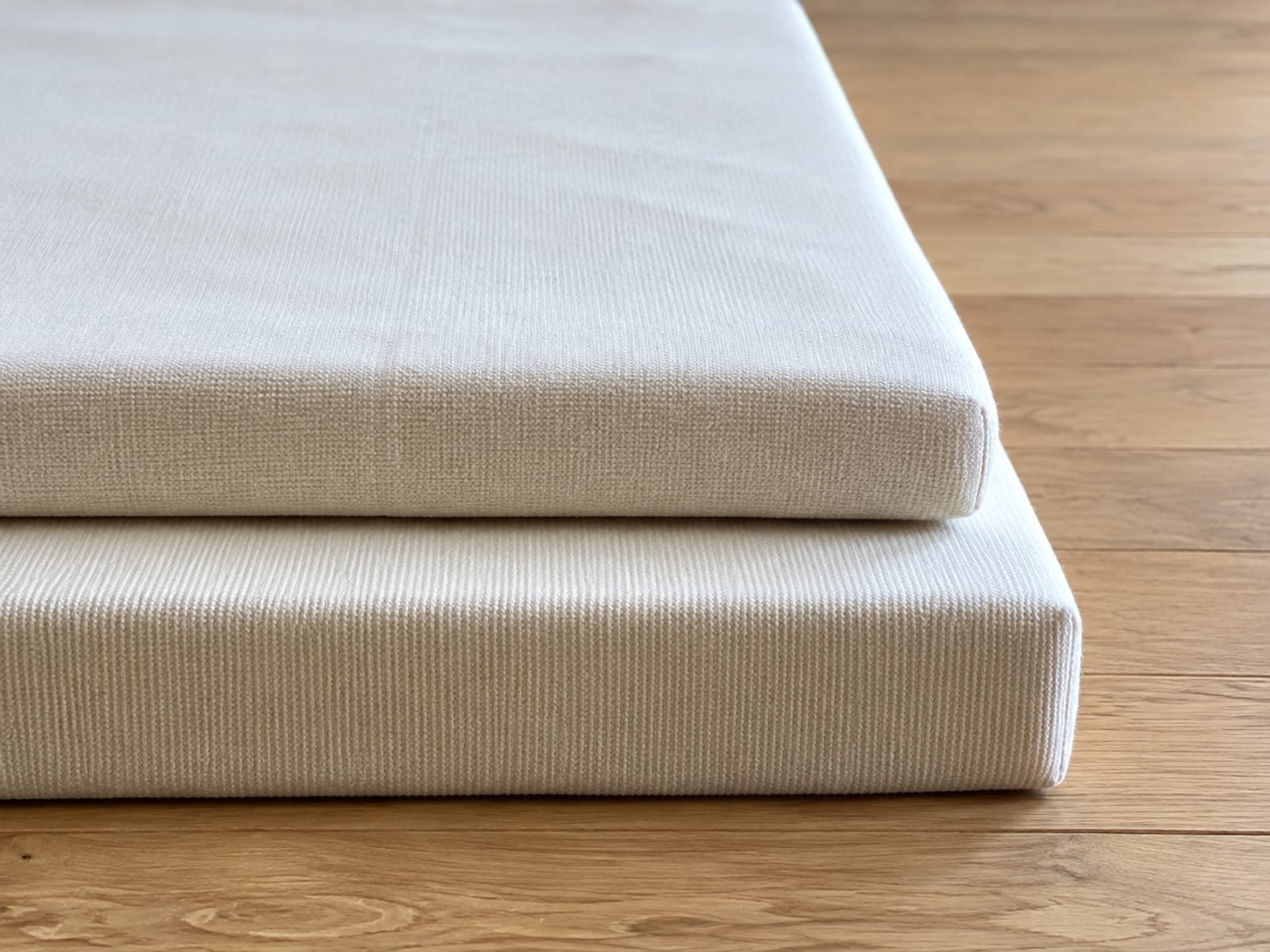 Two plain bare mattress pads of clearly different thickness lying one on the other on bare floorboards, front edges aligned and offset so both cut edges show