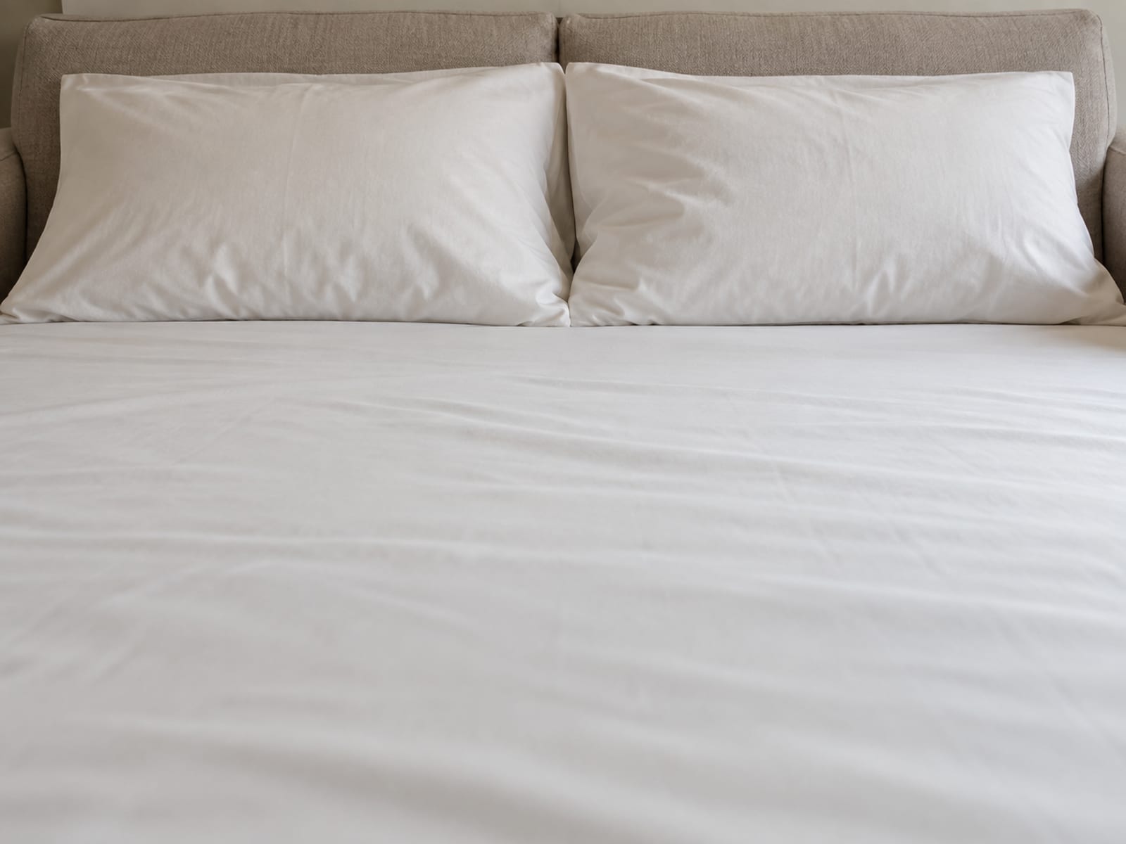 A close view at mattress height across the head end of an opened wide sleeper bed made up with one plain pale fitted sheet, two plain pale pillows lying side by side against the upright back of the sofa, framed so the two pillows and the breadth of sheet they share fill the picture from edge to edge