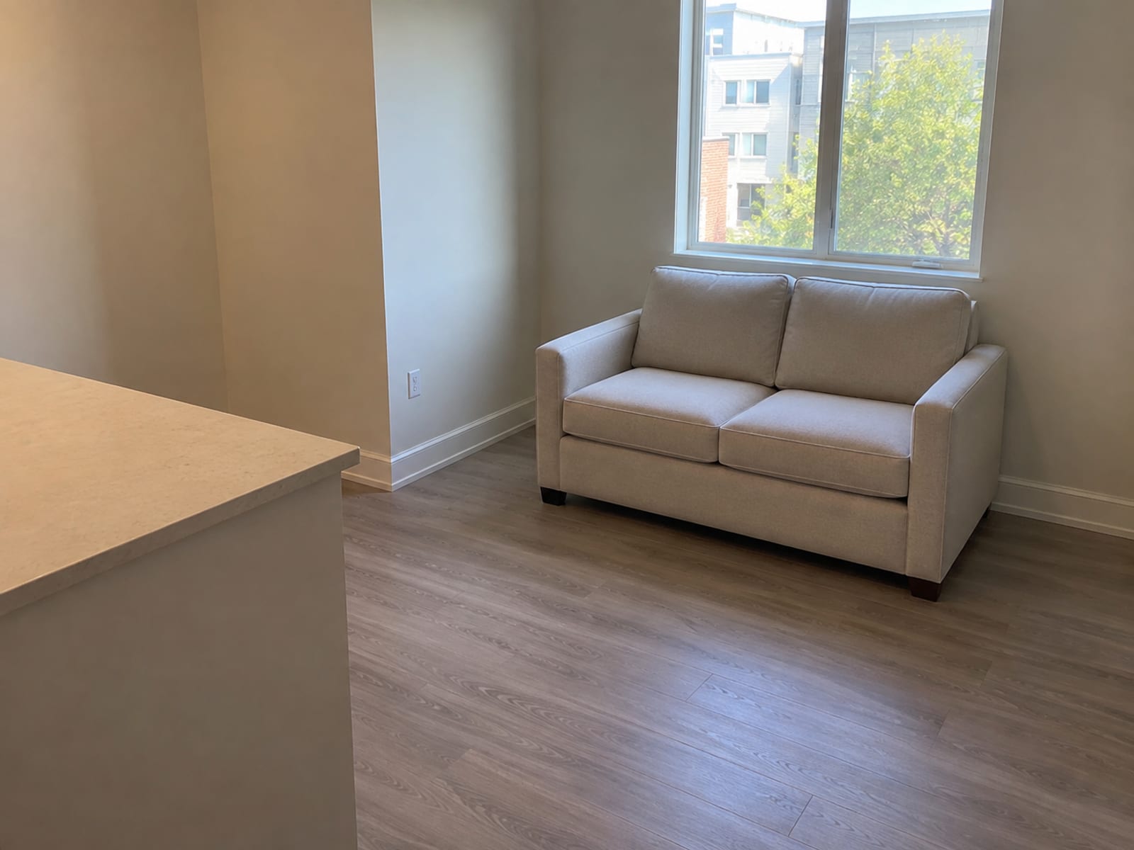 A closed two-seat sleeper loveseat in a pale linen-look weave standing under a window along the one free wall of a small daylit studio apartment, photographed from beside the plain end of a kitchen counter at standing height at a three-quarter angle, the counter's bare edge running along one side of the frame