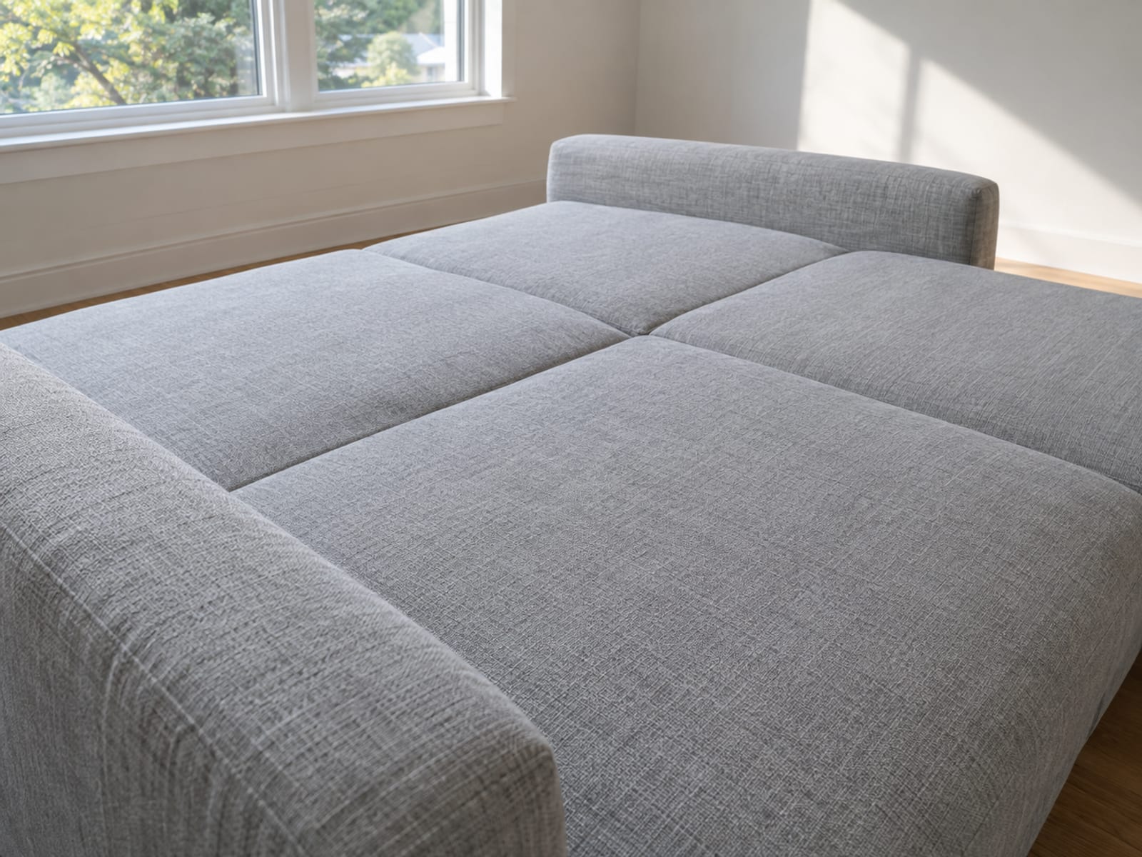 A sofa converted into a bed made only of its own upholstered parts, its pulled-out underframe carrying the sofa's back cushions laid down flat and level with the seat so the whole sleeping surface is a row of plain upholstered sections lying edge to edge, with no separate mattress anywhere, photographed from beside the head end at standing height at a slight downward angle
