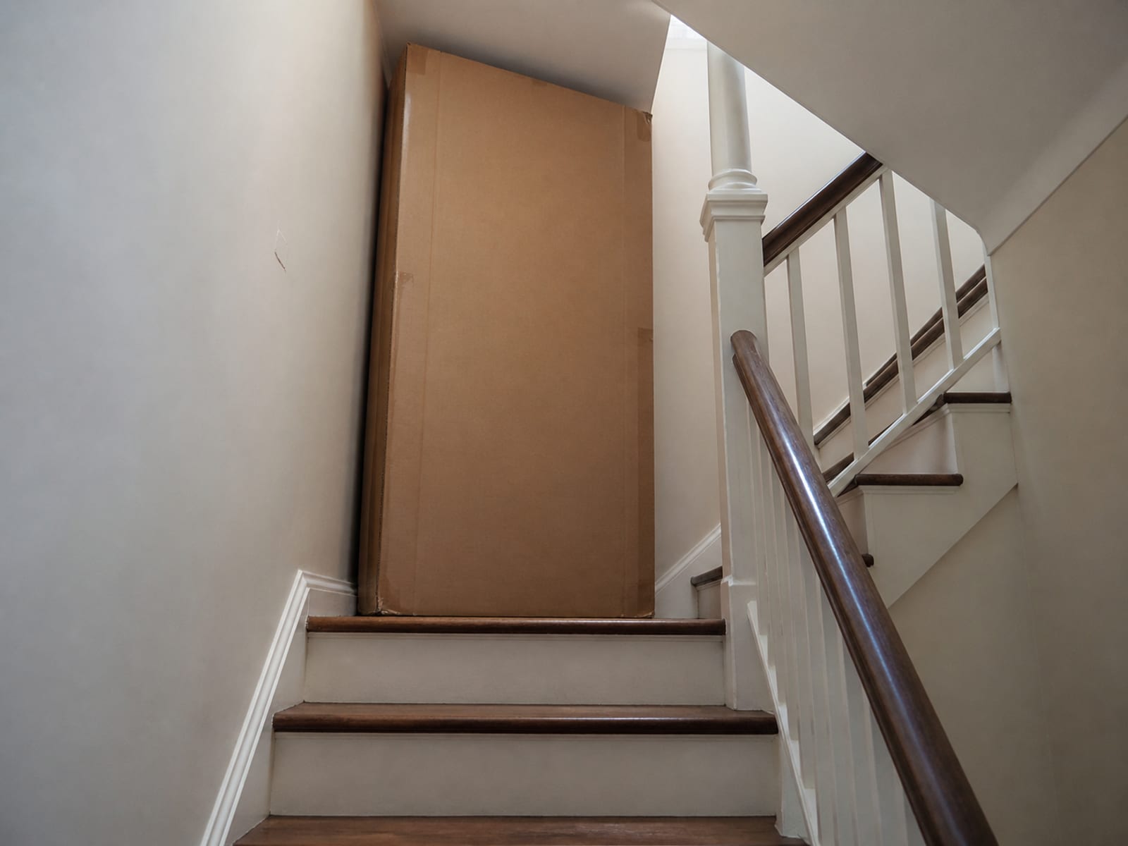 A large flat furniture carton standing on end at the half-landing of a staircase that turns back on itself, with the low ceiling above the turn in frame
