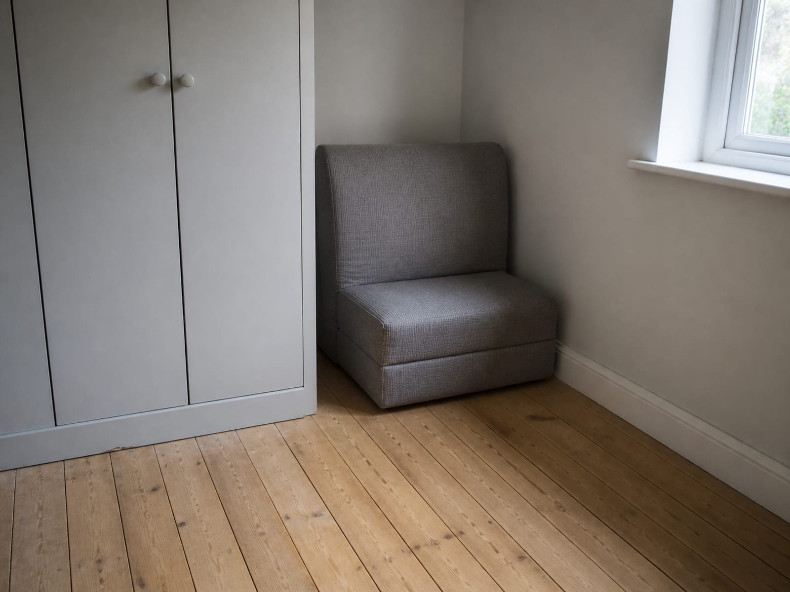 A folded single-seat floor convertible in a corner between a wardrobe and a window, with a narrow strip of walking floor beside it