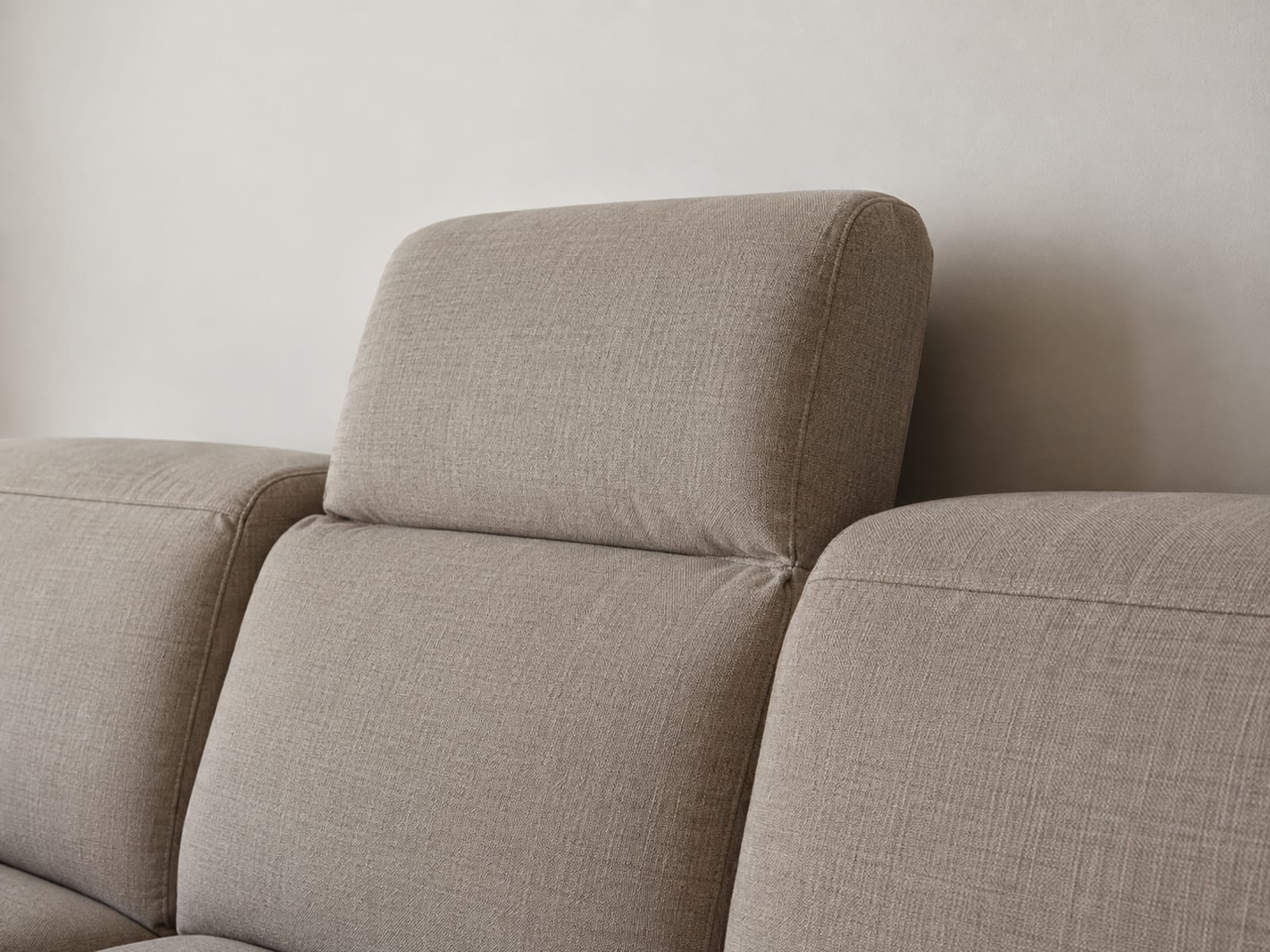 A close side view of the top of one seat's back on an upholstered sofa where a hinged headrest section has been raised and angled forward above the line of the backs on either side of it, framed so the raised headrest and the lower level of its neighbors read together against a plain wall