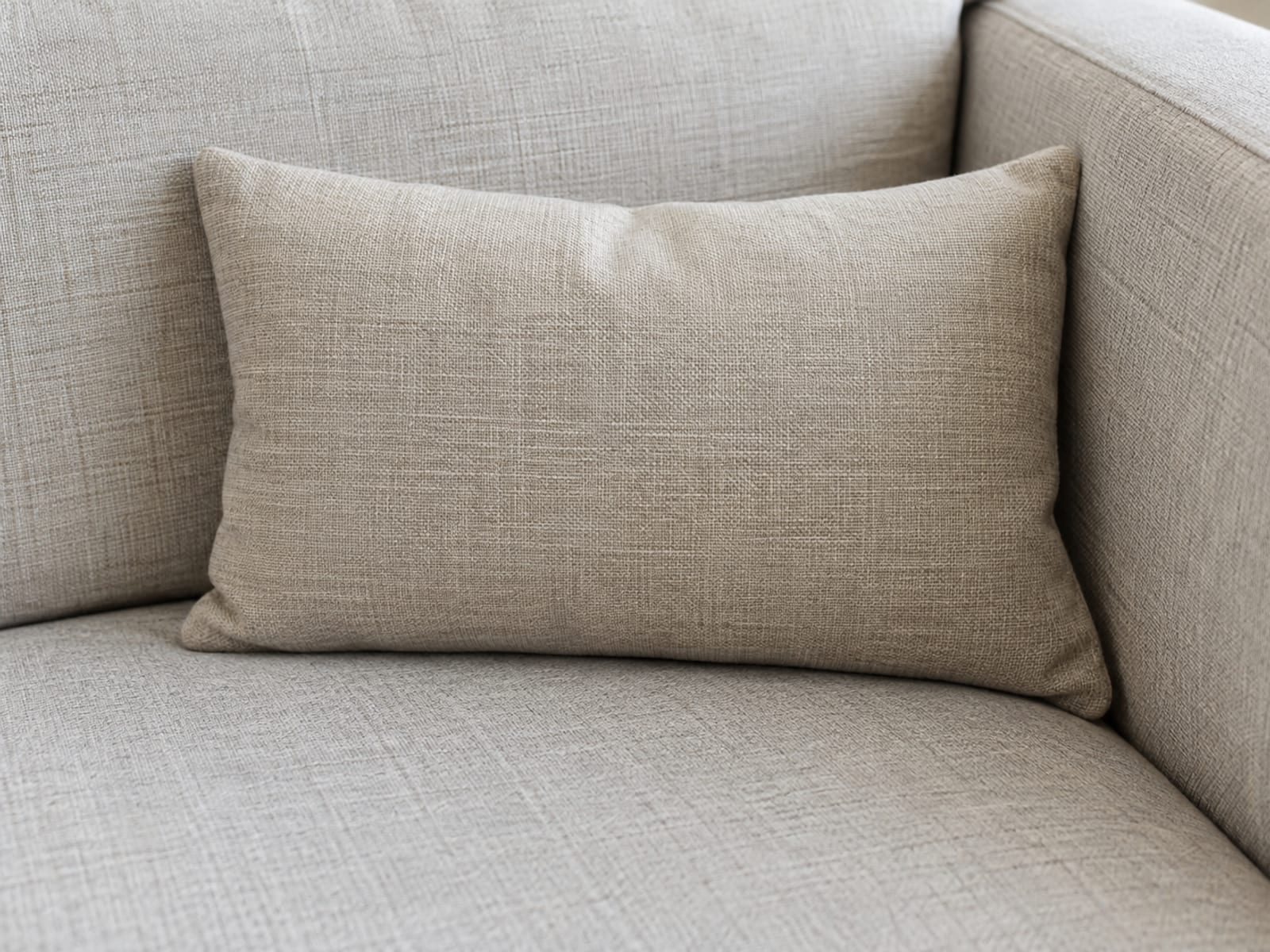 One firm plain rectangular lumbar pillow standing upright in the corner where the back of an upholstered sofa meets its arm, set down low against the base of the back just above the seat, photographed from a front three-quarter angle at seat height