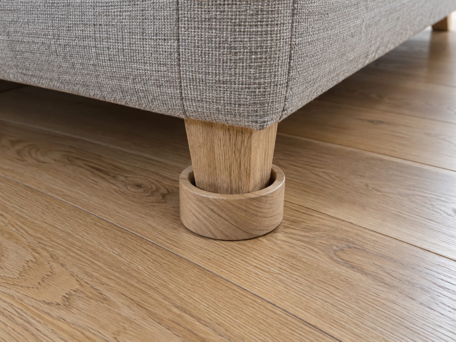 A low close view at floor level of one front corner of an upholstered sofa where its short wooden leg sits squarely in the shallow cup of a plain solid wooden furniture riser standing on bare oak floorboards, framed so the leg, the riser and the lower edge of the upholstery above them fill the picture