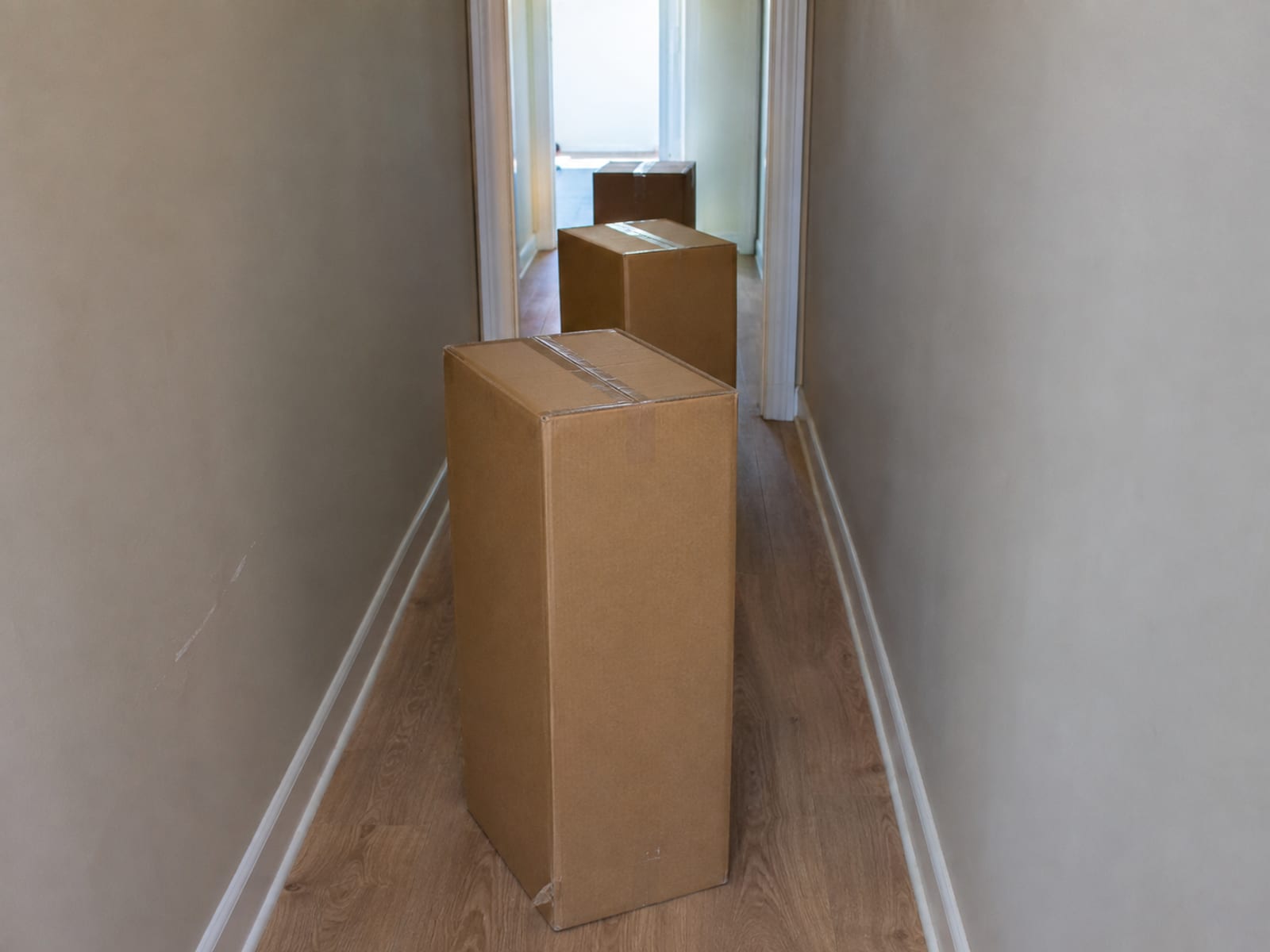 Three large compressed cartons standing one behind another in a narrow hallway, with the doorway they must each pass at the far end
