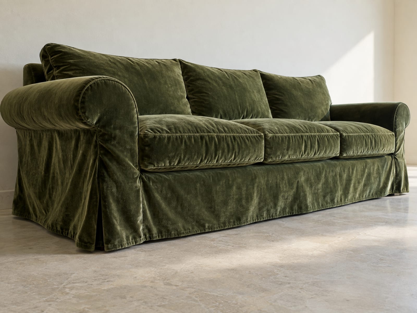 A generic classic rolled-arm sofa with loose seat and back cushions wearing a loose-fit cover in a deep moss velvet, photographed from a low angle along its front from one end in soft side daylight so the velvet's sheen shifts across the relaxed folds at the arm front and along the skirt