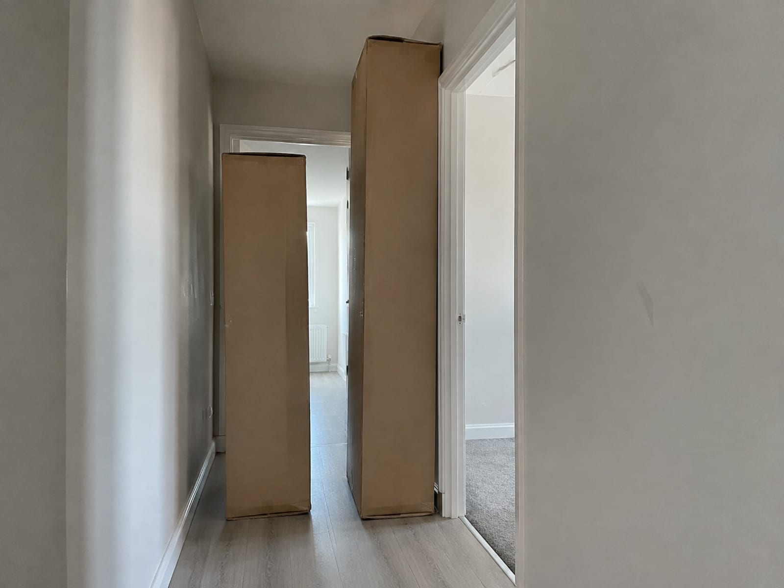 Two tall compressed sofa cartons standing on end in a narrow hallway, one taller than the doorway beside them