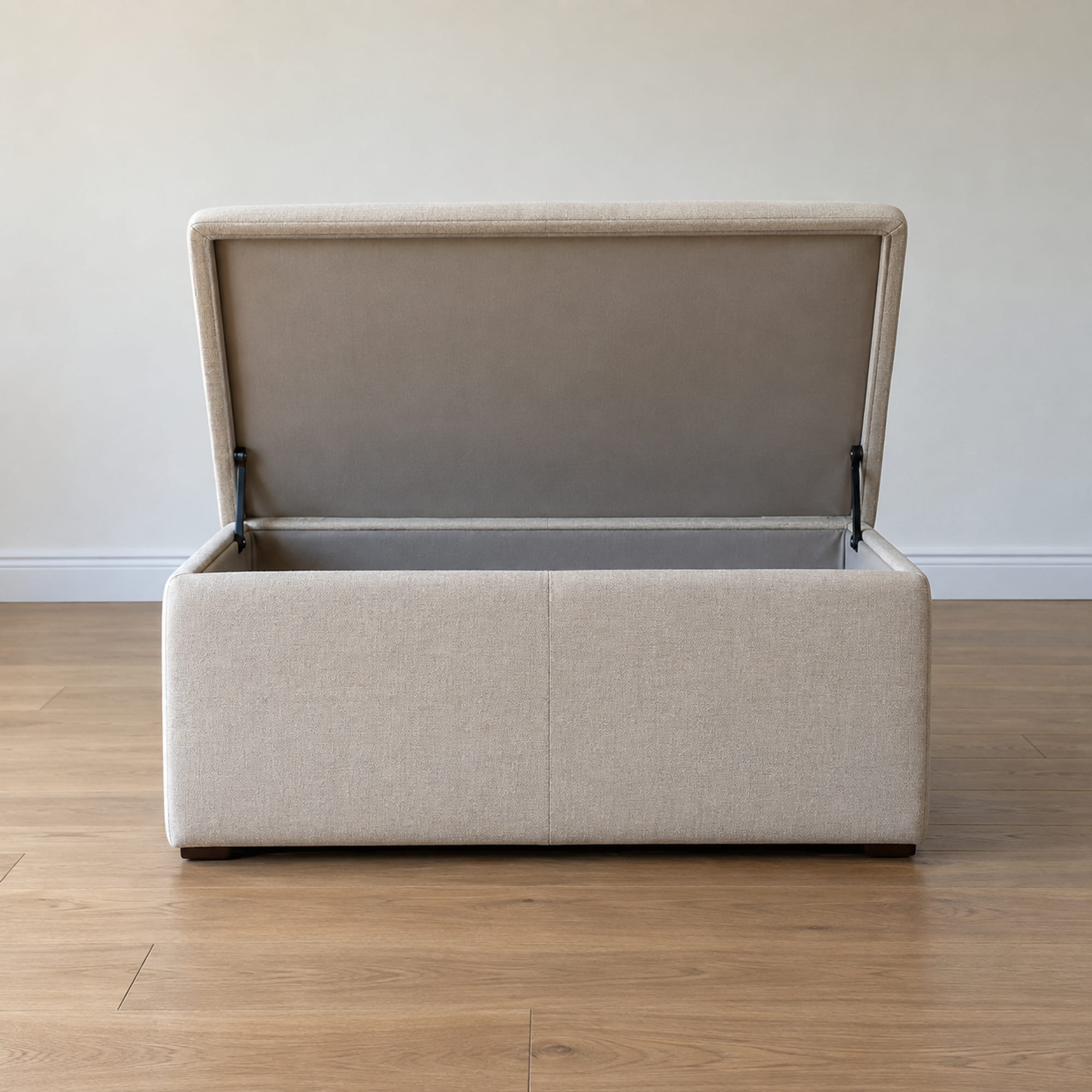 A rectangular upholstered storage ottoman standing on bare oak floorboards in front of a plain wall in an empty daylit room, its hinged lid standing open and upright at the back so the lid's padded top faces away and its plain lined underside faces the camera as an upright panel behind the open box, photographed square on from the front at a low height close to the ottoman's own top edge