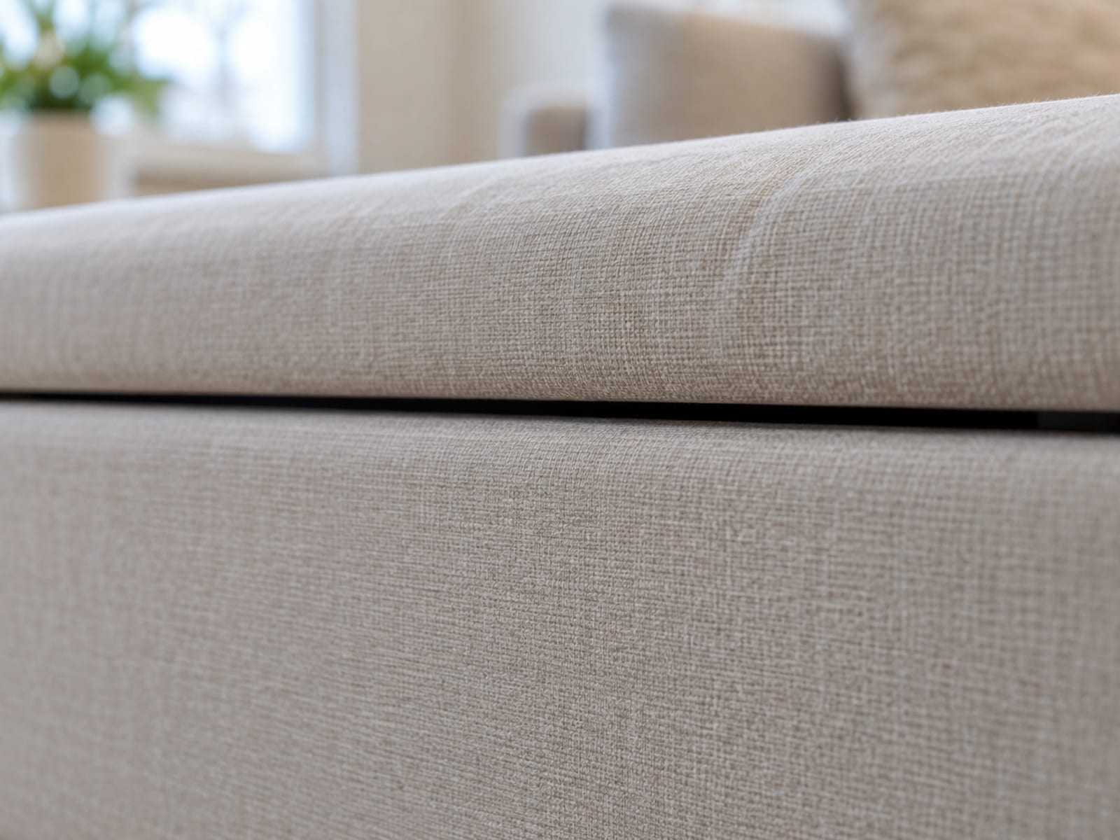 A low close side view along the front edge of a closed storage ottoman where the upholstered lid rests on small plain bumpers so a narrow continuous gap runs between the underside of the lid and the rim of the box, the dark line of the gap running across the frame as the plain subject