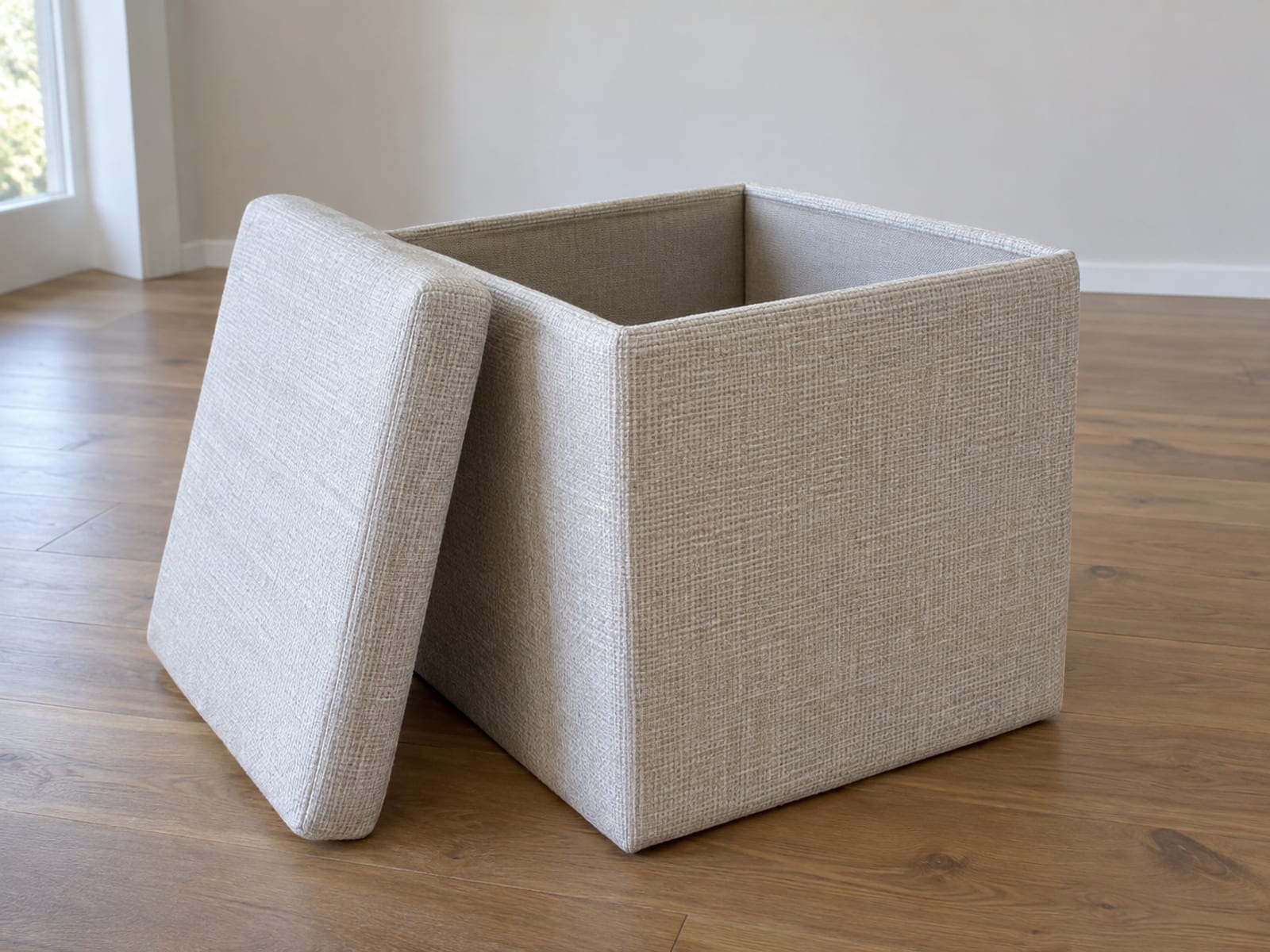 A square upholstered storage ottoman with its loose lift-off lid removed and stood on its edge on the floor, leaning against one side of the ottoman, photographed from standing height at a front three-quarter angle so the lid's thickness and the open square box beside it read as two separate parts