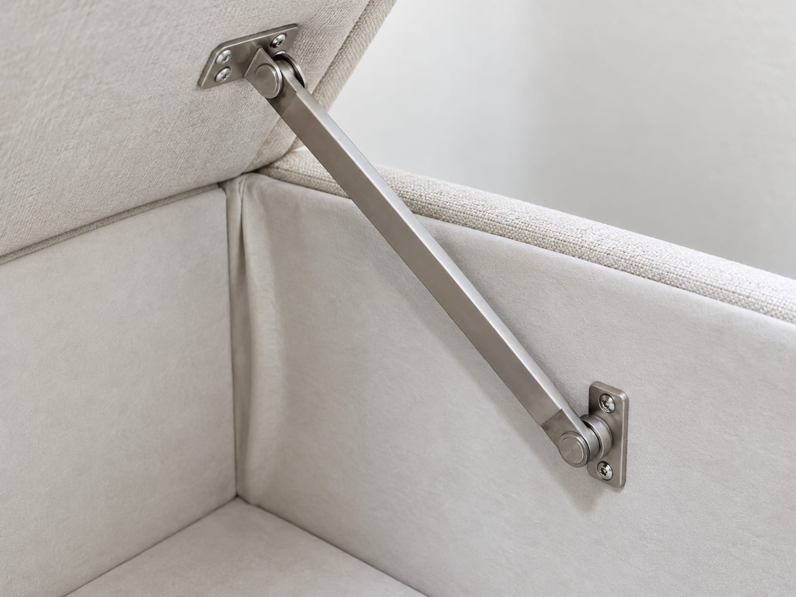 An extremely close view of the inside back corner of an open storage ottoman, where a plain metal slow-close stay arm runs from the underside of the raised lid down to the side wall of the box, framed so the arm and its two mounting points fill the picture with the lined interior around them