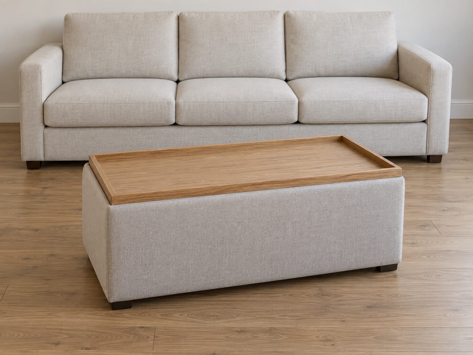 A rectangular upholstered storage ottoman standing in front of a plain sofa in a daylit living room, with its reversible lid turned over so a flat plain oak tray face sits upward on top of the box, photographed from standing height at a slight downward three-quarter angle